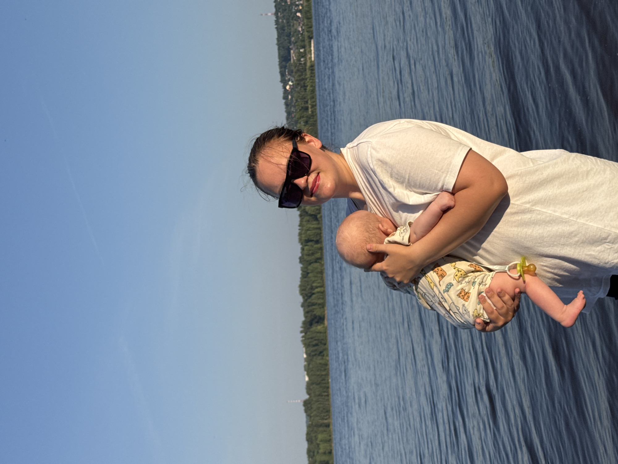 In Noviy, Russia, a woman wearing sunglasses and a white t-shirt stands by a body of water, holding a baby. The baby is swaddled in a light-colored, patterned garment and has a yellow pacifier clipped to it. The background shows a dense line of green trees along the opposite shore, with a town and a tower visible in the distance under a clear blue sky. The water's surface is rippled, suggesting a gentle breeze.