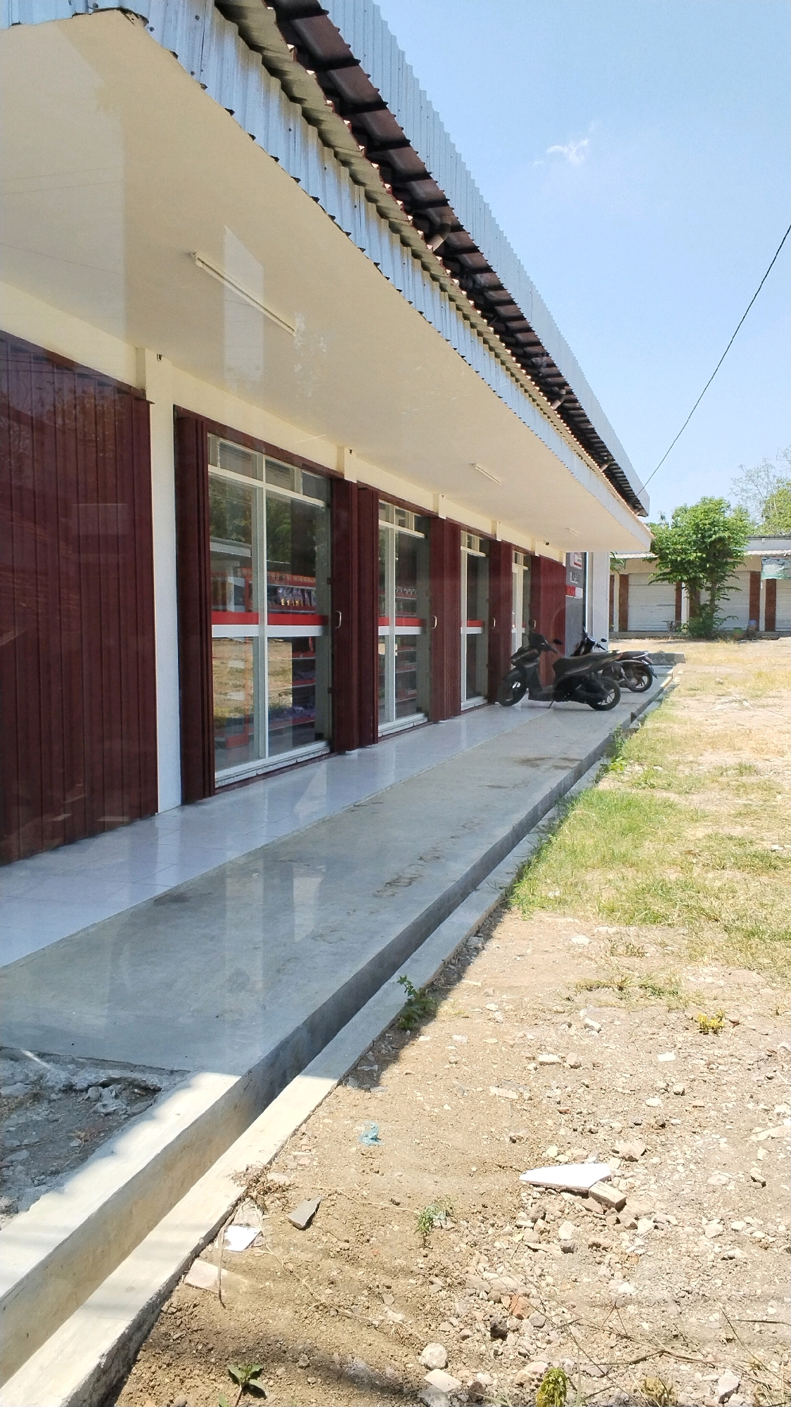 The image depicts the exterior of a row of shops with brown metal roller doors and large glass windows, located on a sunny day in Pulorejo, Indonesia. A concrete walkway runs in front of the shops, bordered by a drainage ditch. Two motorcycles are parked on the paved area in front of the shops. The ground outside the walkway is dirt and sparse dry grass. In the background, there are more buildings with corrugated metal roofs and trees. The sky is clear blue with a few wispy clouds.