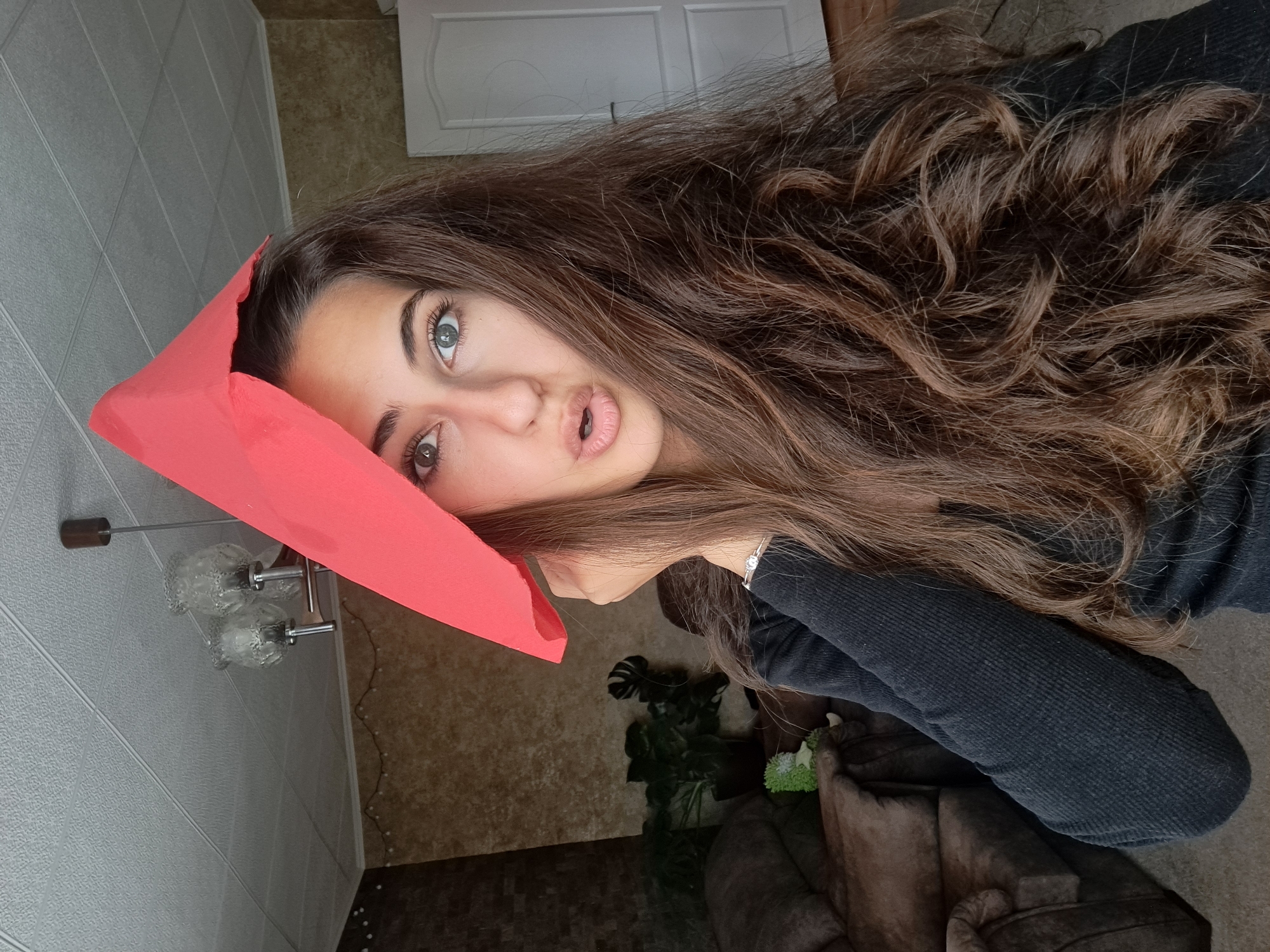 A young woman is captured in a close-up portrait, with the image rotated 90 degrees clockwise from its likely original orientation. When correctly oriented, she is looking directly at the camera with her head tilted slightly to her right. She has long, voluminous, wavy brown hair that cascades over her shoulders. Her eyes are light-colored, appearing to be green or grey-blue, and she has dark, well-defined eyebrows. Her lips are slightly parted, forming a soft "o" shape.
A bright reddish-pink piece of folded paper or thin fabric rests on the left side of her forehead, partially obscuring her hair and extending down past her left eye. She is wearing a dark, possibly black or navy blue, ribbed long-sleeved top. A silver-colored bracelet with small shiny details is visible on her left wrist, which is positioned near her chin, seemingly supporting her head.
The setting appears to be indoors. Behind her head and to her left (when correctly oriented), a wall with light-colored, textured paneling is visible, along with a white panel door in the upper left corner. A wall-mounted light fixture with two glass shades and chrome accents is also visible on this wall. To her right and behind her, the wall has a patterned, possibly stone or mosaic-effect, brown wallpaper. A dark brown, plush armchair or sofa is partially visible in the lower right, and a potted plant with large green leaves (resembling a Monstera) is situated between the woman and the furniture. The flooring in the bottom left corner appears to be light brown tiles. The woman is actively posing for the photograph. There is no visible text.