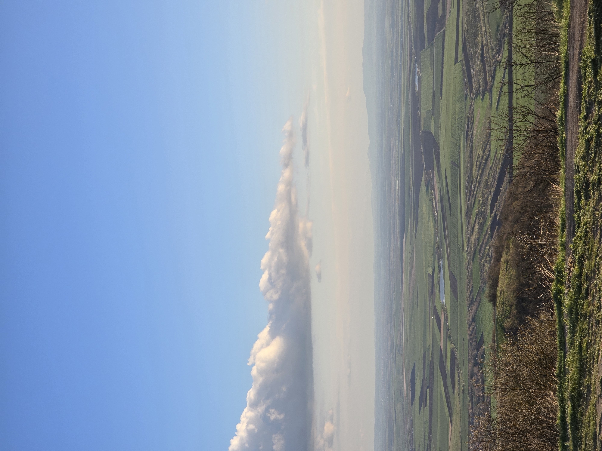 The image depicts a landscape near Pobeda, Bulgaria. The sky is predominantly blue with some white clouds. The foreground shows green fields and vegetation, with a dirt road running along the edge. Some trees and bushes are also visible.