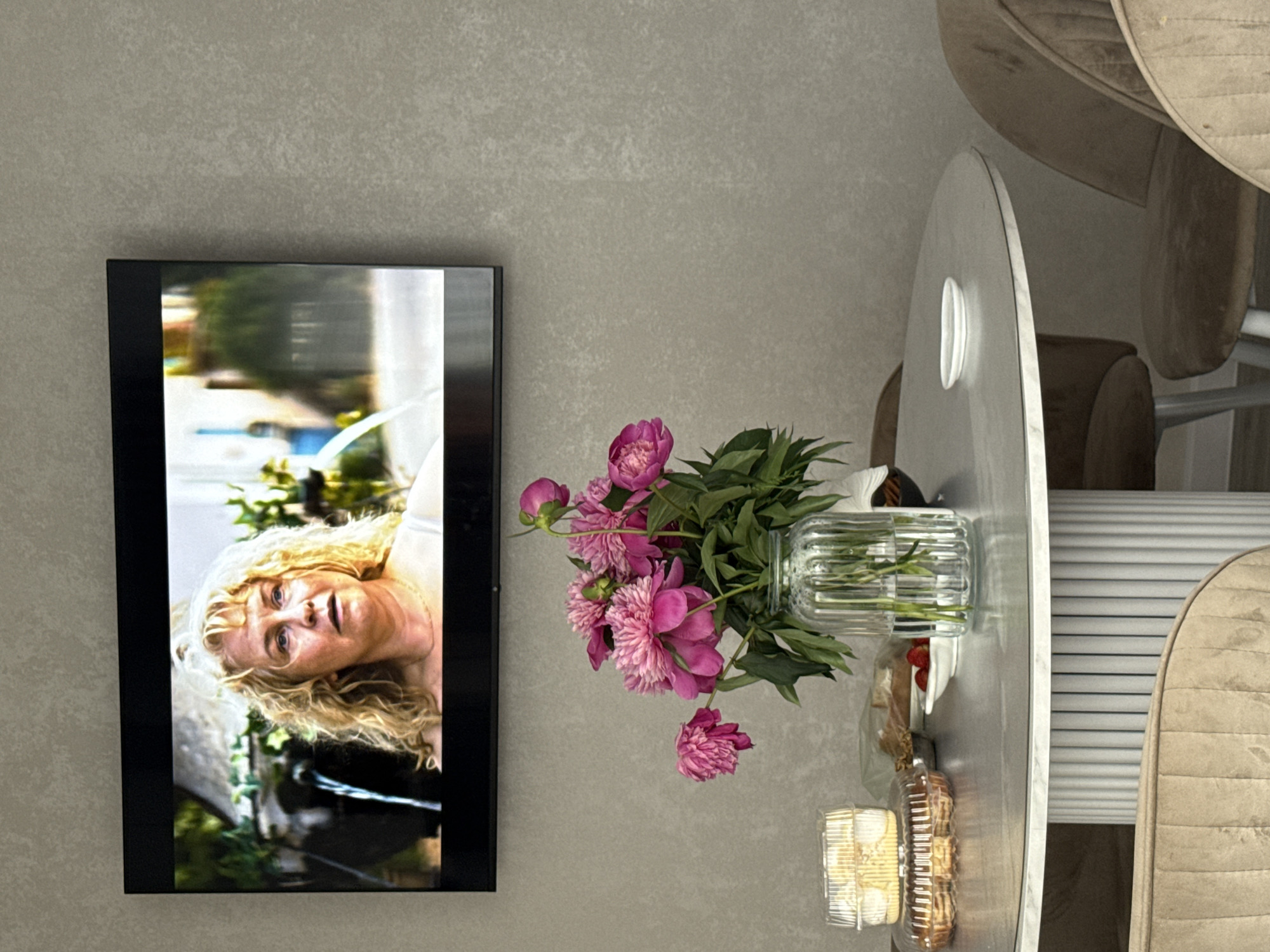 A television screen displays a close-up of a woman with blonde, curly hair and a concerned expression. Below the television, a round dining table holds a vase of pink peonies, several containers of pastries, a small bowl of strawberries, and a tiny white dish. Two plush, taupe-colored chairs are partially visible on either side of the table. The wall behind the television and table has a textured, grey finish. The scene appears to be indoors, likely in a dining or living area. The location is specified as Nizhnyarkeyevo, Russia.