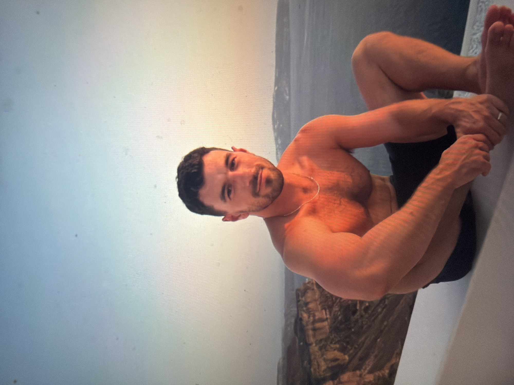 A male subject is depicted seated on a white, angled ledge, overlooking a panoramic coastal landscape during what appears to be the golden hour of sunrise or sunset. The location is specified as the city of Shumen, Bulgaria.
The man has short, dark hair and a neatly trimmed beard or stubble. He is shirtless, revealing a muscular build, and wears dark swim shorts. A thin silver chain necklace is visible around his neck, and a ring is present on the ring finger of his left hand. He looks directly at the camera with a slight smile. He sits with his knees bent, his left hand resting on his right ankle area, and his right arm resting on his right knee. His skin is illuminated by the warm, orange-red glow of the setting or rising sun.
The background features a vast expanse of calm, dark blue water (likely a sea or ocean) stretching to the horizon. To the left, rugged, reddish-brown cliffs descend sharply towards the water. In the far distance on the right, hazy landforms or islands are visible across the water. The sky transitions from a light blue at the top to a soft, warm orange and yellow glow near the horizon, indicative of a clear atmosphere. The immediate surface below the man is a smooth, clean white ledge.