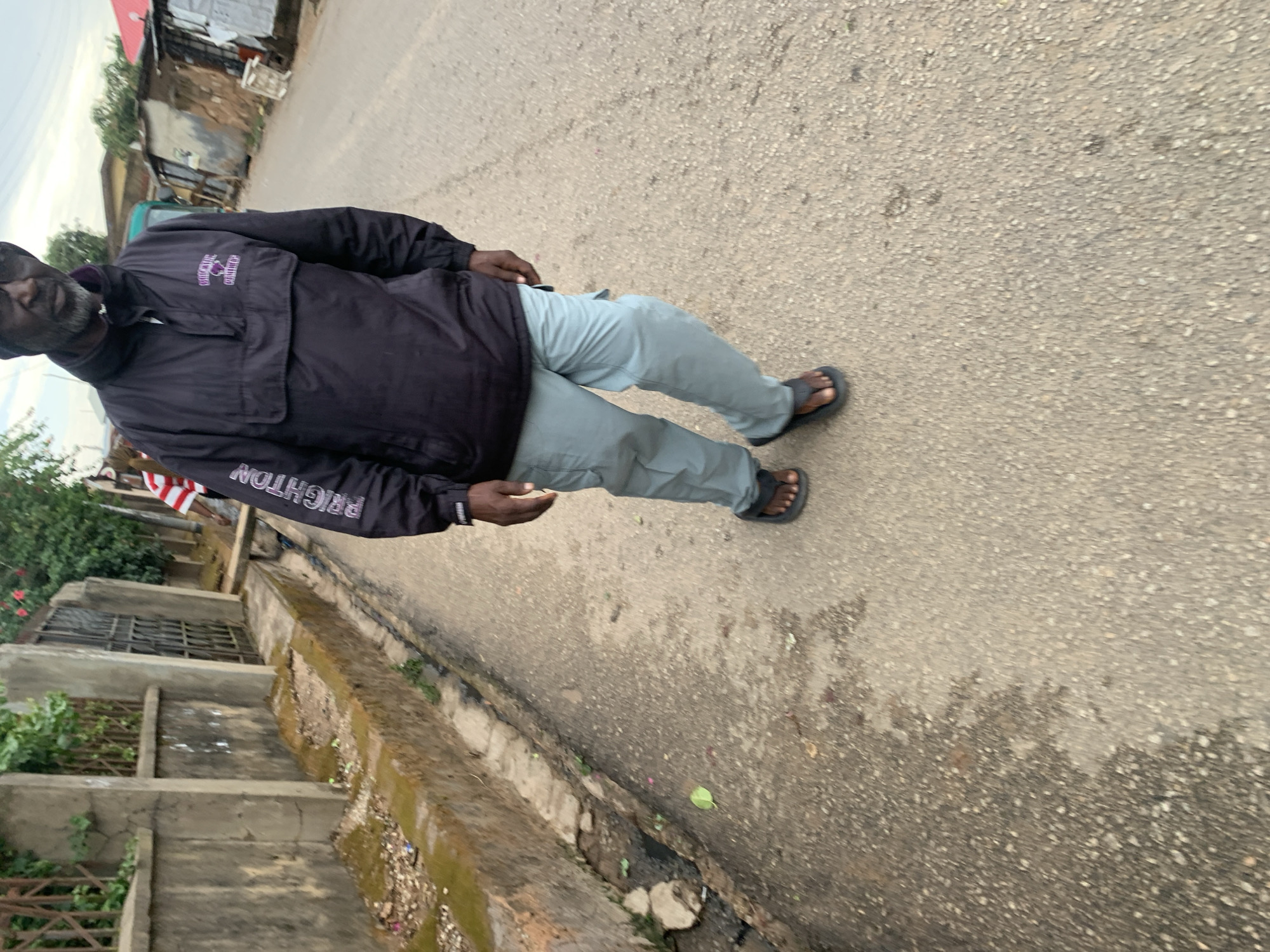 The image depicts an older, dark-skinned man walking on a paved road. He wears a dark jacket with "BRIGHTON" printed on the sleeve, light-colored pants, and sandals. The road appears to be within the city of Jos, Nigeria, based on the style of the architecture and infrastructure in the background. A concrete drainage ditch runs along the edge of the road, with some vegetation growing in and around it. Buildings and trees are visible in the background.