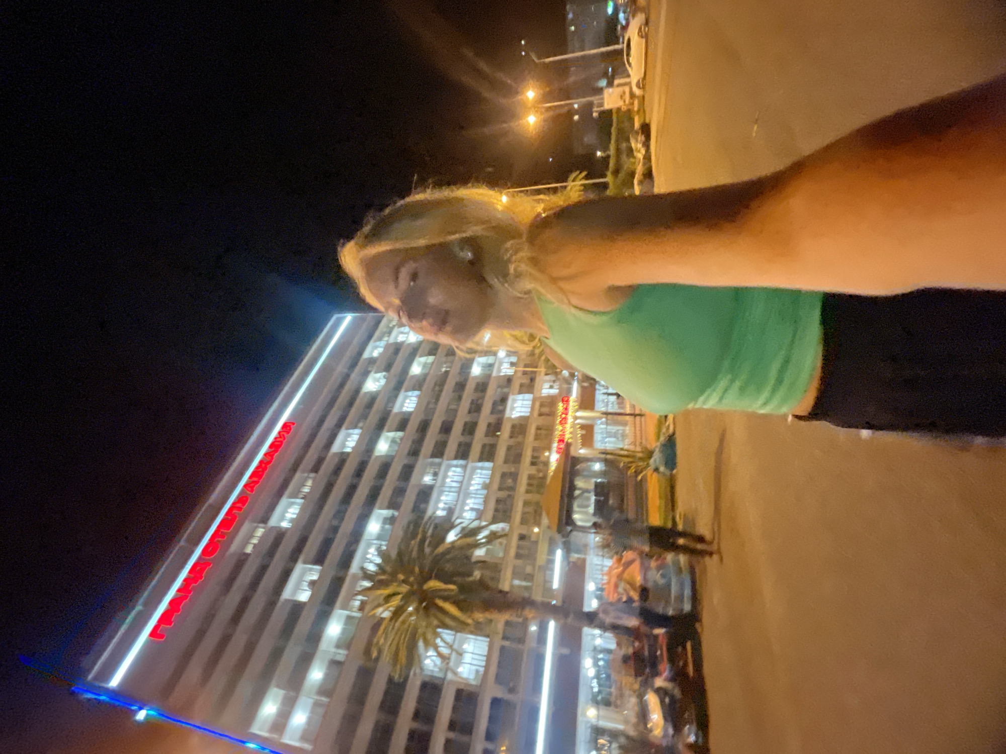 The image captures a night scene in Gagra, Abkhazia, featuring a woman in the foreground taking a selfie. She is wearing a green sleeveless top and dark pants. Behind her, a large, modern building is illuminated with red neon lights that spell out "ГРАНД ОТЕЛЬ АБХАЗИЯ" (Grand Hotel Abkhazia). A palm tree stands in front of the hotel, and several people, vehicles, and streetlights are visible in the background, indicating a public area or street. The overall atmosphere suggests a well-lit urban environment at night.