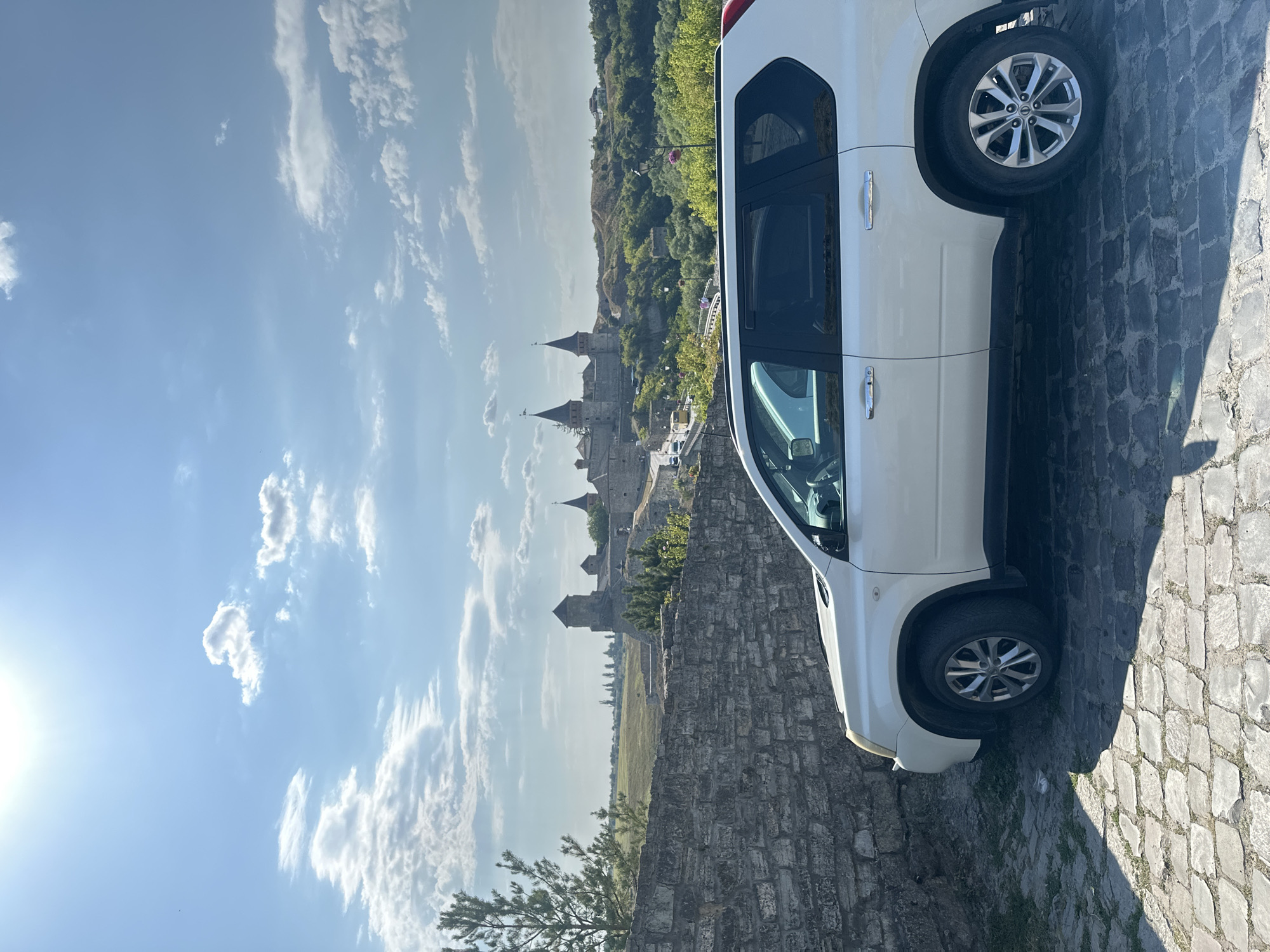 A white SUV is parked on a cobblestone street in front of a stone wall. In the background, the Kamianets-Podilskyi Castle in Ukraine is visible on a hill under a blue sky with scattered clouds. The sun is shining brightly. The scene is a daytime, outdoor view.