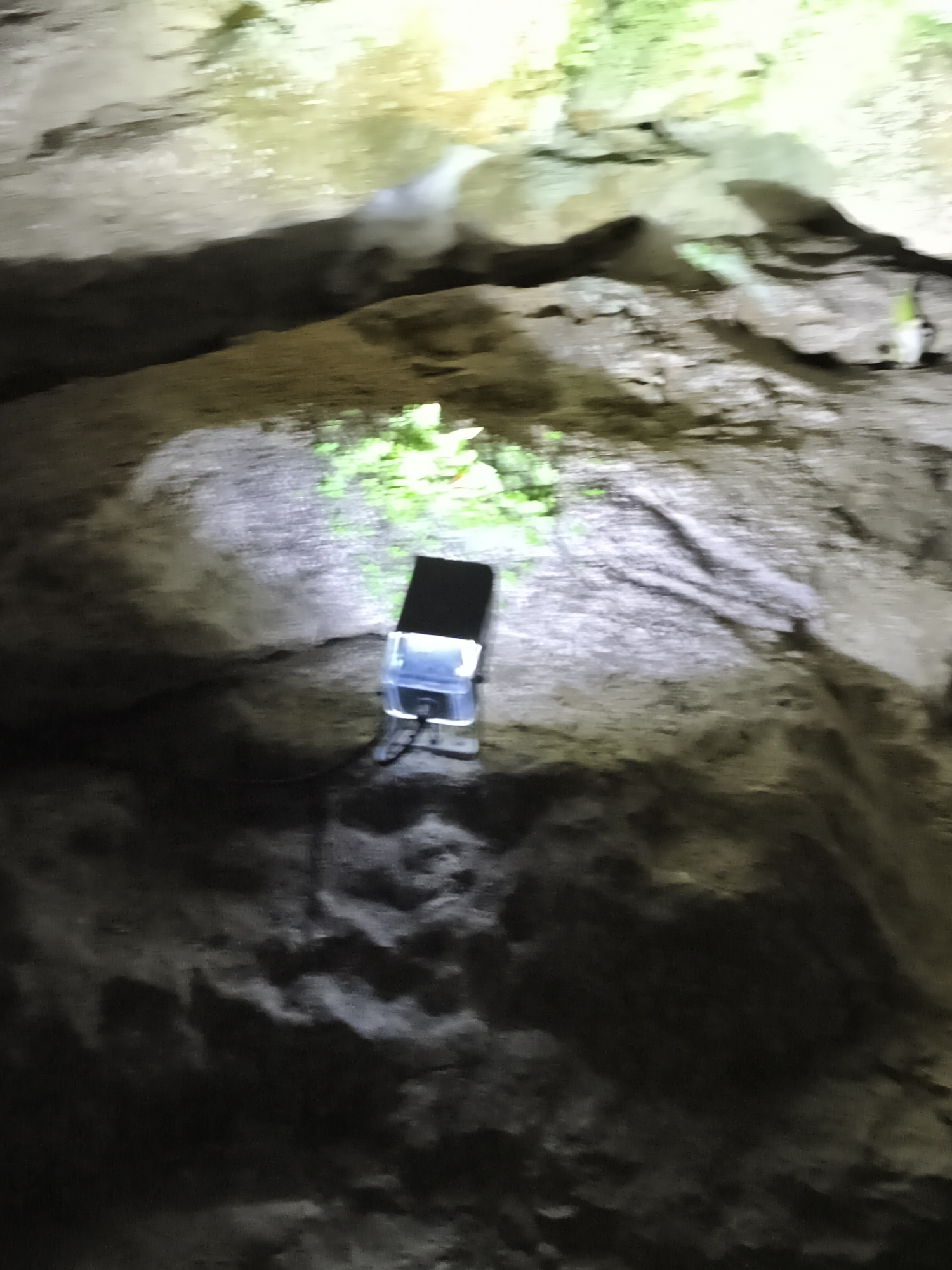 A black object with a clear plastic cover and a black top is placed on a textured, rocky surface. The object is illuminated by a bright light, casting shadows. In the background, more textured rocks are visible, with some areas lit by a green light source. The overall scene appears to be within a cave or an outdoor rocky environment. This image was captured at the Balinger Felsen, a natural rock formation located in Hayingen, Germany.