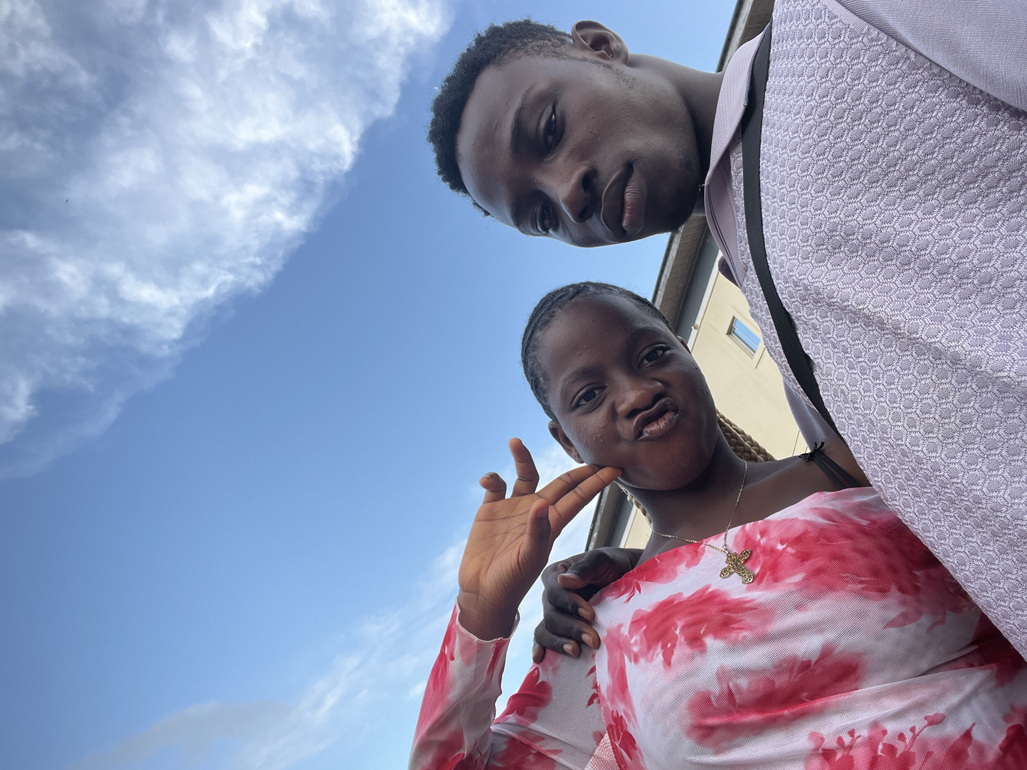 In Nkpor, Nigeria, this selfie captures two young people with clear blue skies and scattered clouds as the backdrop. The male subject, on the upper right, is angled downwards, his face partially visible with a thoughtful expression. He is wearing a light purple patterned shirt over a dark strap. The female subject, positioned in the lower center, is looking directly at the camera with her lips pursed and one hand touching her cheek, fingers splayed in a gesture. She wears a sheer white top with a pink tie-dye pattern and a gold cross necklace. The image is framed by a building on the right and vast open sky filling the rest of the frame.