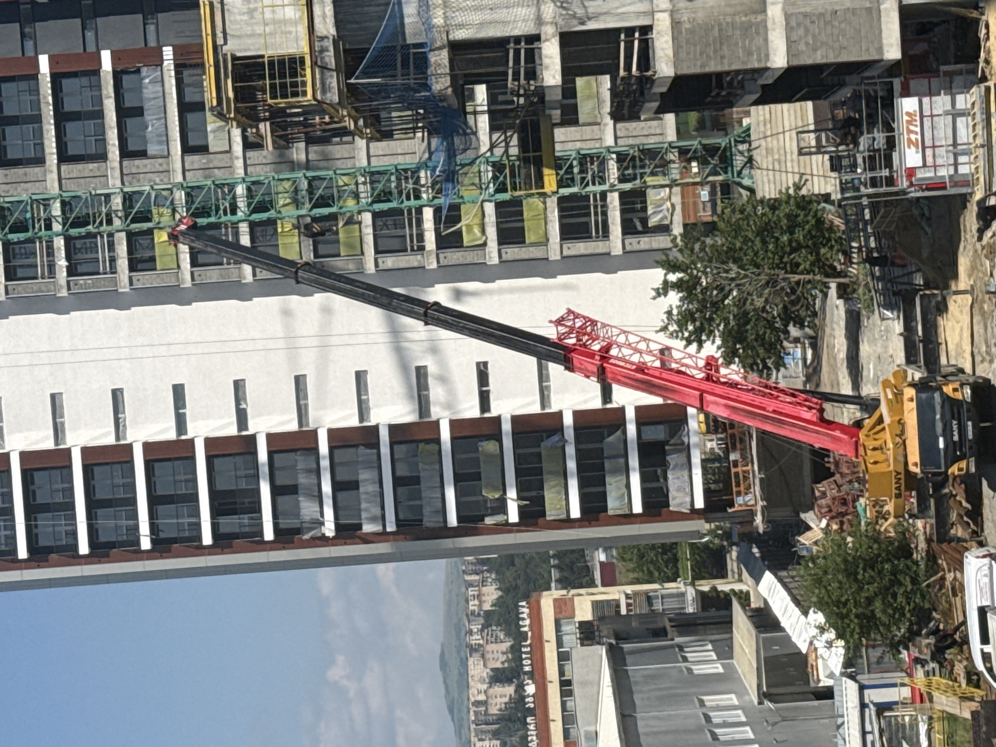 The image captures a construction site in Tbilisi, Georgia, with a focus on a tall building under construction and a large crane. The main building is characterized by its modern architecture, with multiple stories featuring rectangular windows and balconies. To the left of this building, another structure is visible, appearing to be a hotel with the signage "HOTEL AGAVA" prominently displayed. In the background, a cityscape with hills and numerous buildings suggests an urban environment.
A prominent feature of the scene is a substantial red and black crane, its long boom extending diagonally across the frame, reaching towards the upper floors of the construction site. The crane is positioned next to a green lattice-like structure, likely a part of the building's permanent crane system. Below the crane, a yellow truck with "SANY" branding is visible, suggesting it's part of the construction equipment.
The construction site itself is a hive of activity, with various building materials scattered around, including wooden pallets and what appear to be stacks of bricks or concrete blocks. Some parts of the building are unfinished, with exposed concrete and scaffolding. Safety netting is also visible, indicating ongoing construction work. A few trees, one with dense foliage, dot the landscape, adding a touch of greenery amidst the concrete and steel. The sky is blue with some clouds, indicating daylight. The overall impression is that of a bustling urban development project in progress.