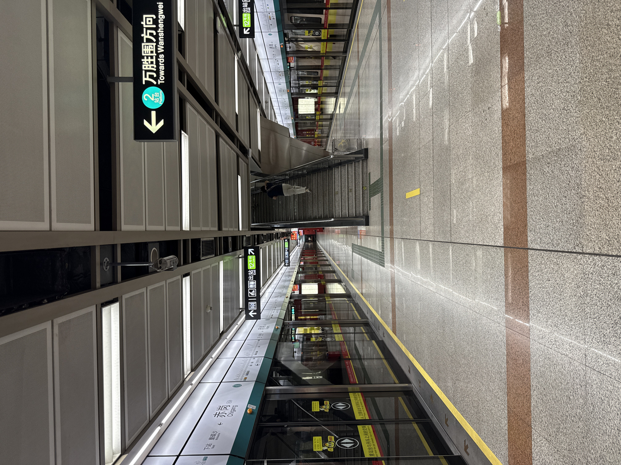 The image depicts the interior of a subway station, specifically Platform 2, indicated by signage reading "2 站台 万胜围方向" and "Platform 2 Towards Wanshengwei". The station features a modern design with a high ceiling and fluorescent lighting. The platform is lined with automated doors that separate it from the subway tracks. A train with red and silver carriages is visible at the platform. The floor is tiled with a speckled gray pattern, marked with yellow and reddish-brown stripes. Signage in both Chinese and English is present throughout the station, providing directions and information. A single person is seen walking up a set of stairs on the right side of the image. The environment appears clean and well-maintained, characteristic of an operational subway system.