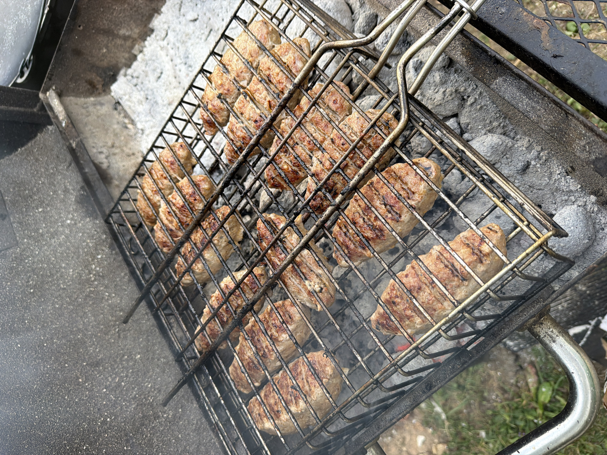 This image depicts an outdoor barbecue scene. A grill basket filled with rows of grilled meat is positioned over a bed of gray ashes, suggesting an active cooking process. The meat appears to be in the form of elongated, oval-shaped patties, with visible grill marks indicating they have been cooked on both sides. Smoke is rising from the grill, further emphasizing that the food is being cooked. The background shows the rough, textured surface of the barbecue's lid, as well as some greenery and soil to the left, indicating an outdoor setting. The scene is devoid of any people or specific landmarks that would identify its precise location within Bayreuth, Germany.