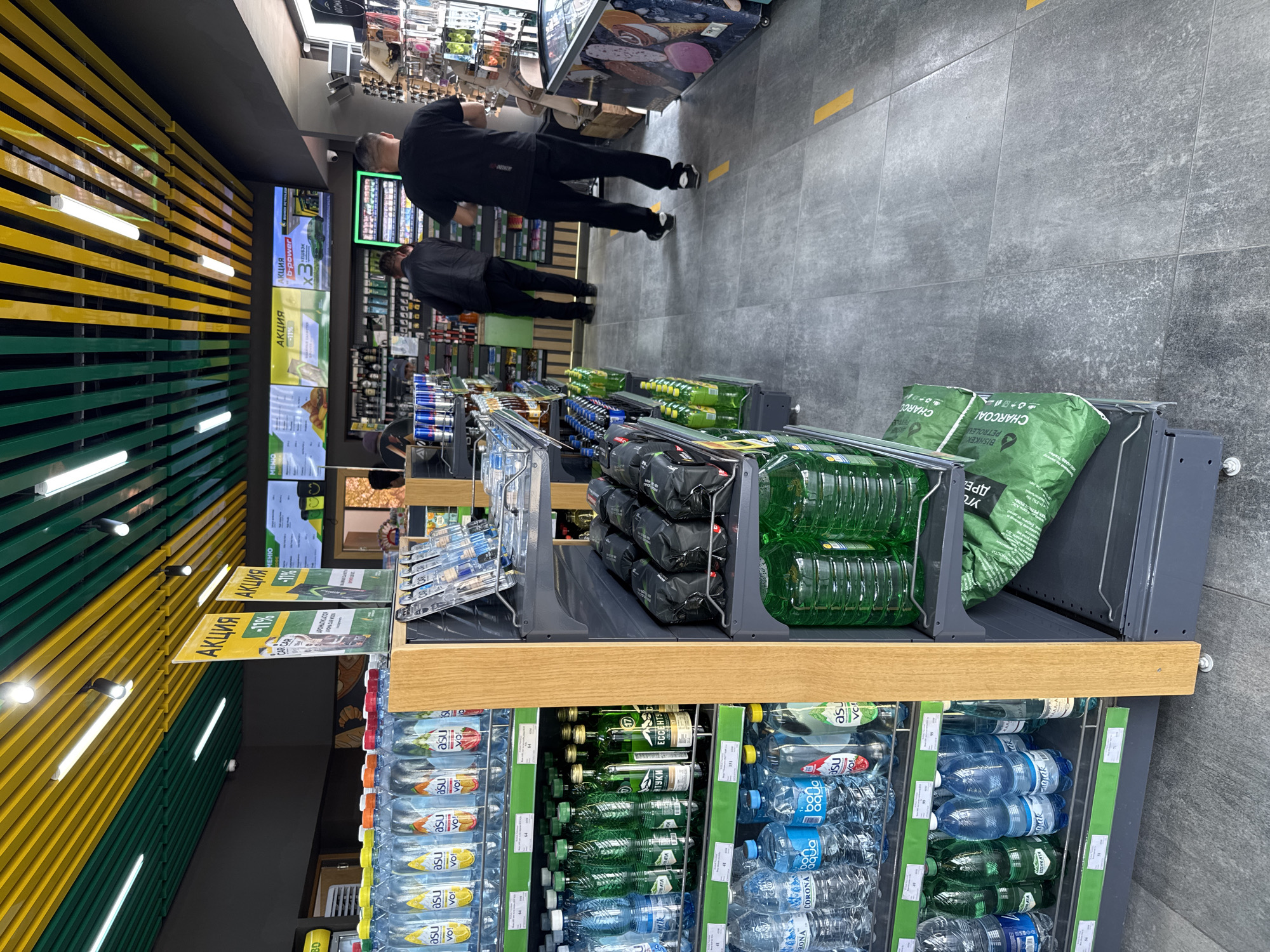 The photo shows a grocery store aisle with shelves stocked with various items, including bottled water, charcoal, and other goods. Two men are walking down the aisle. The walls are decorated with yellow and green stripes. The store appears to be modern with gray tiled floors.
Several signs marked "АКЦИЯ" in cyrillic appear on the shelves, suggesting a sale. The presence of brands with cyrillic labels indicates the location is likely in a country where Russian is spoken or understood.
The inclusion of brands such as "CORONA" and the fact that Kyrgyzstan labels include products in English suggest this store is in Кашат, Kyrgyzstan.