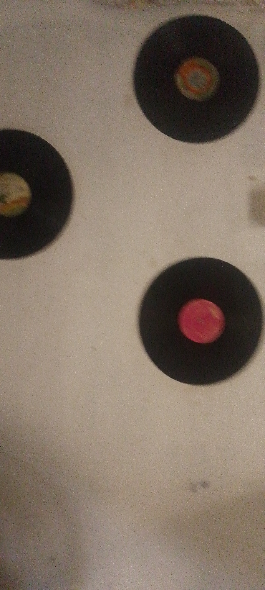 Three vinyl records are arranged on a plain, light-colored surface. The records are black with a central label. The top record has a label with concentric circles of orange, yellow, and blue. The left record has a label that is mostly light-colored with some indistinct colored markings. The bottom record has a solid pink central label. The scene is likely indoors, given the lighting and background. There are no people or actions depicted.