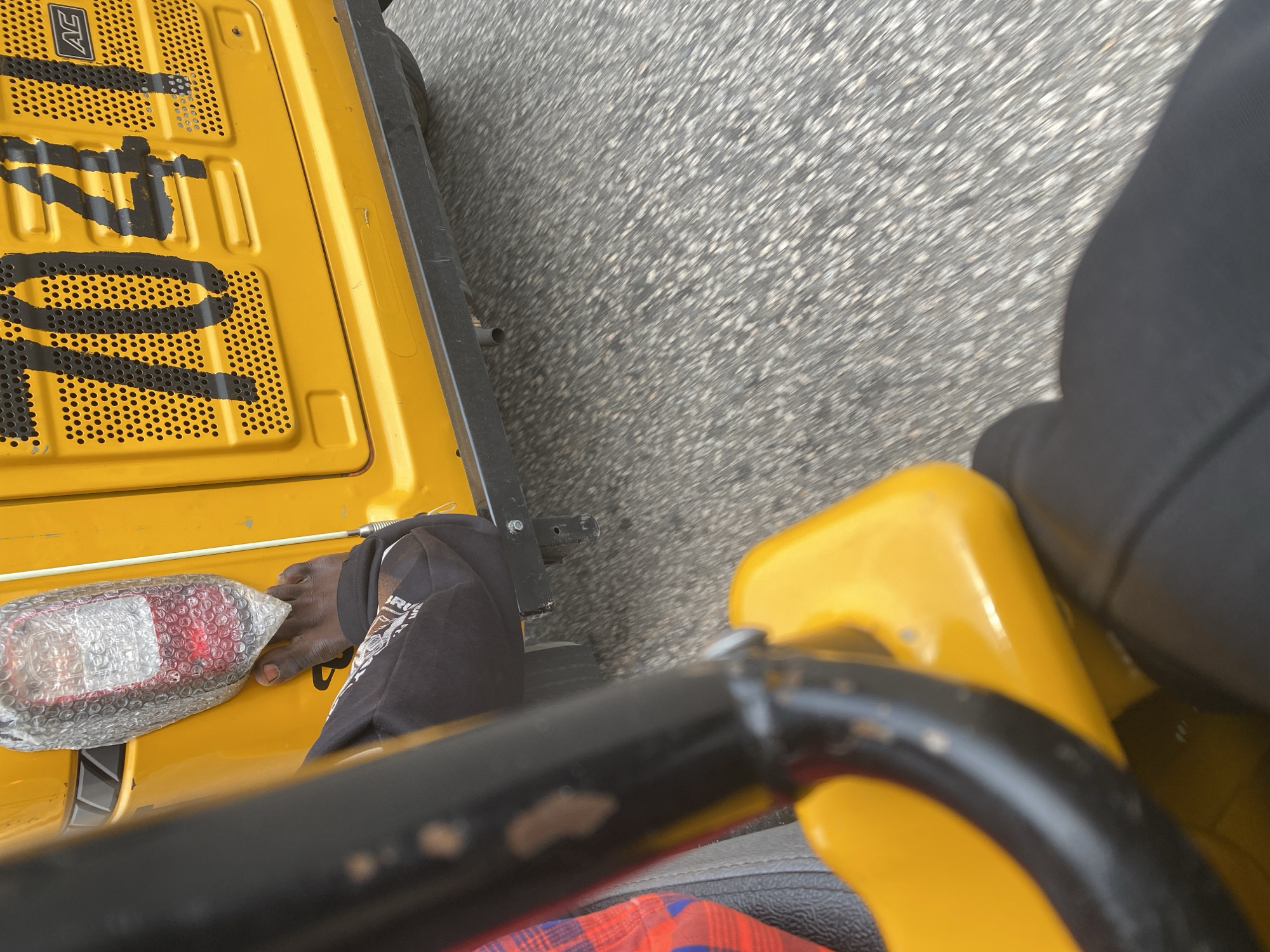 The image shows a first-person perspective from a moving yellow vehicle, likely a three-wheeled auto-rickshaw, on a paved road. The ground, covered in asphalt with white and gray speckles, is blurred due to motion. The vehicle's yellow body is visible, along with a black metal bar in the foreground, suggesting a handle or part of the structure.
A person's feet, wearing black pants or shorts, are visible near a red taillight wrapped in bubble wrap. The taillight is mounted on the yellow body of the vehicle. The person's bare foot is positioned near the taillight, with toes pointed slightly.
On the yellow surface of the vehicle, there are large, black, stencil-like numbers and letters, partially visible and smudged. One segment clearly shows the letters "AC" within a rectangle. The numbers appear to be "1770" or similar. The overall impression is one of a functional vehicle, possibly in use for transportation or delivery. The bright sunlight suggests it is daytime, and the weather appears clear, enabling outdoor travel. The setting appears to be an urban or semi-urban environment given the paved road and markings on the vehicle. No specific emotional cues are discernible from the provided view.