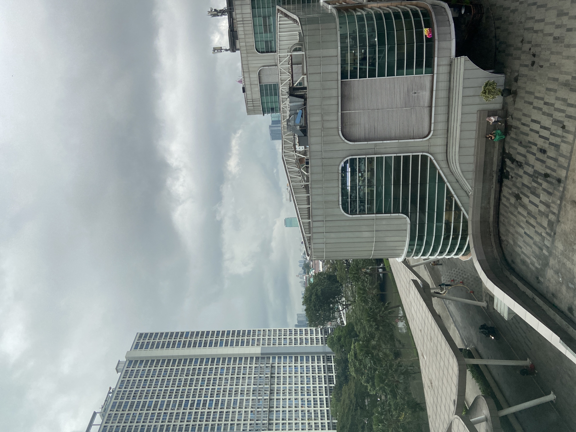 The image shows a cityscape in North Jakarta, Indonesia, taken from an elevated position. Prominently featured is a modern building with large windows and white curved architectural elements. To the right of this building, a pedestrian walkway with a geometric pavement design is visible, along with a few people sitting near a tree. In the distance, a tall, rectangular building with a grid-like facade of numerous windows is observed. The sky is overcast with grey clouds. The scene contains a mix of urban architecture and greenery with the park and trees.