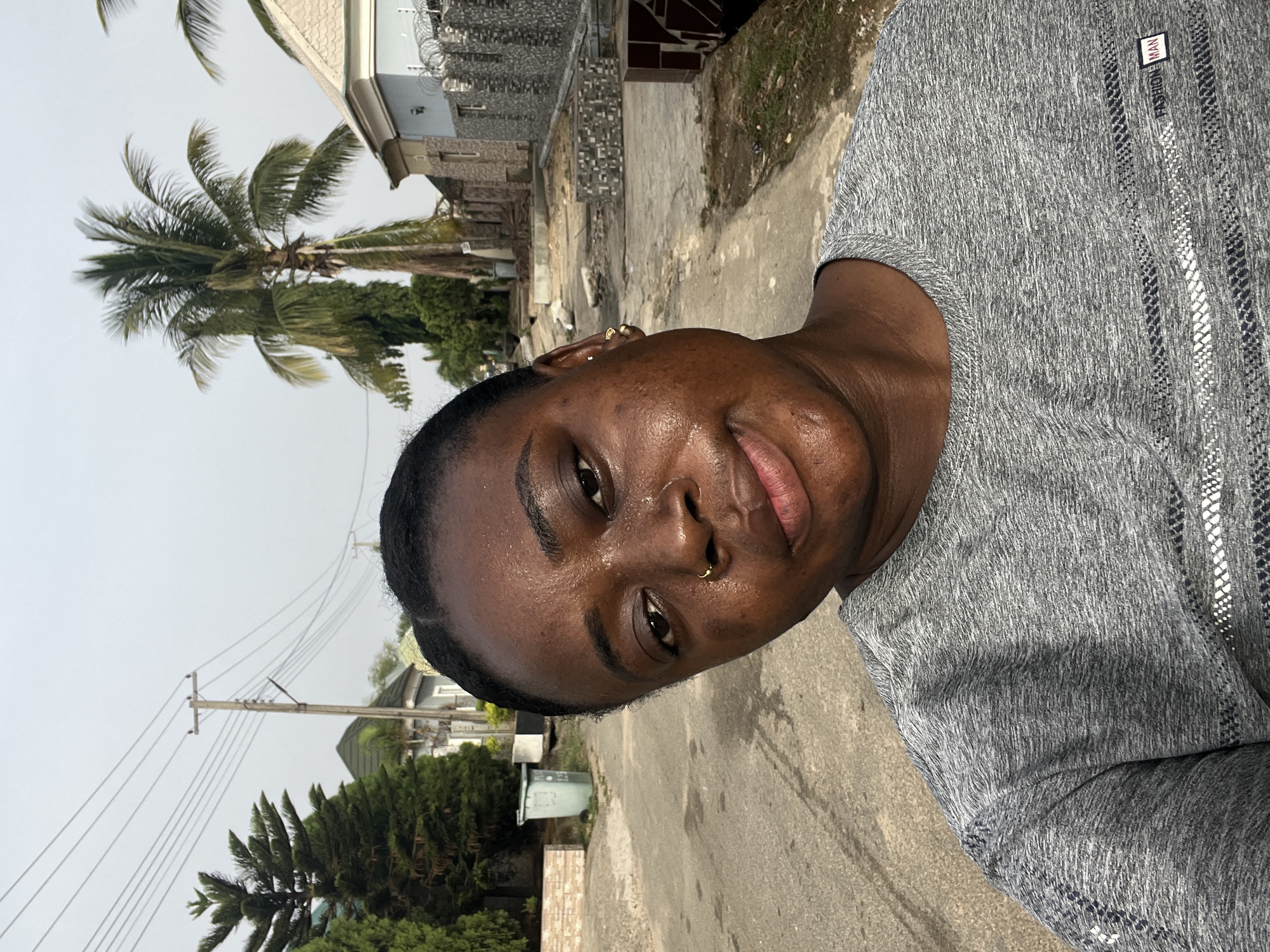 A woman with dark skin and short hair stands on a paved road, likely in a residential area in Abuja, Nigeria. She is wearing a grey t-shirt and has a nose ring. A palm tree and other trees are visible, along with houses and utility poles.