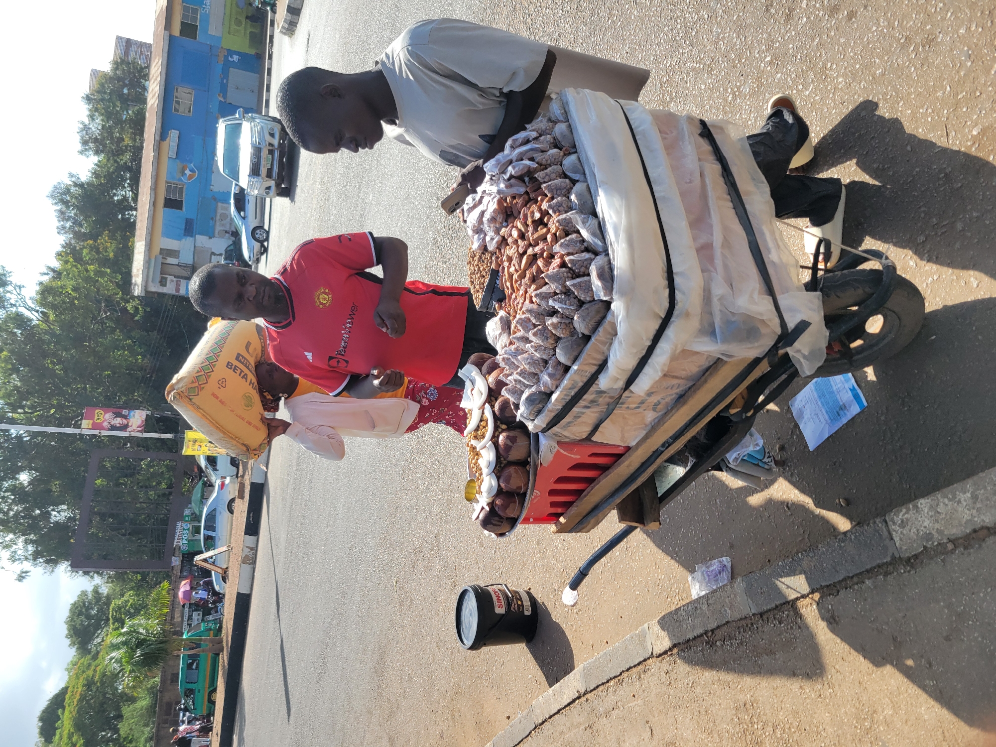 In Barakin Lerawi, Nigeria, two young men and a woman are seen on a roadside. The woman, partially visible, carries a large sack of grain on her head. The man in the red shirt, likely a vendor, stands by a cart filled with various goods. He is wearing a Manchester United jersey and black trousers. To his right, another young man, dressed in a grey t-shirt and dark trousers, holds a smartphone and appears to be interacting with the items on the cart. The cart itself is laden with food items, including nuts and what appear to be dates or similar dried fruits, packaged in small plastic bags and larger containers. A black bucket with a label "SINOP" is positioned to the left of the cart. The scene unfolds on a paved road with a concrete curb, suggesting an urban or semi-urban environment. Vehicles, including a white SUV and a green van, are visible in the background, along with buildings and trees. Shadows indicate a bright sunny day.