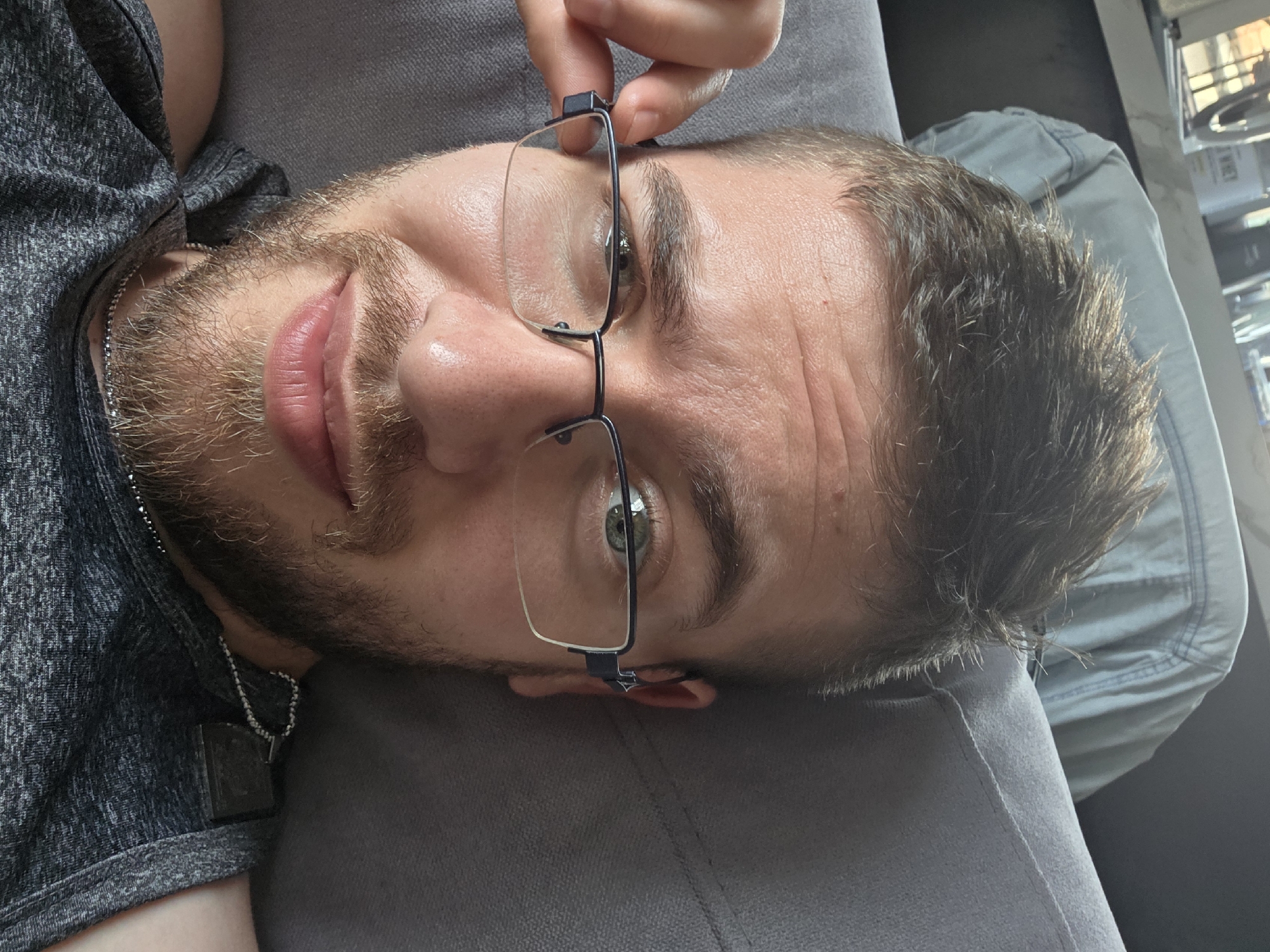A close-up, slightly tilted image shows a man lying down, possibly on a gray sofa or similar surface. He is fair-skinned with light brown hair and a beard. He is holding a pair of eyeglasses in his hand, near his forehead. He is wearing a dark gray shirt and a silver chain necklace with a pendant. The background is blurry, showing a glimpse of what appears to be the inside of a room with daylight coming in. The location is not discernible from the image.