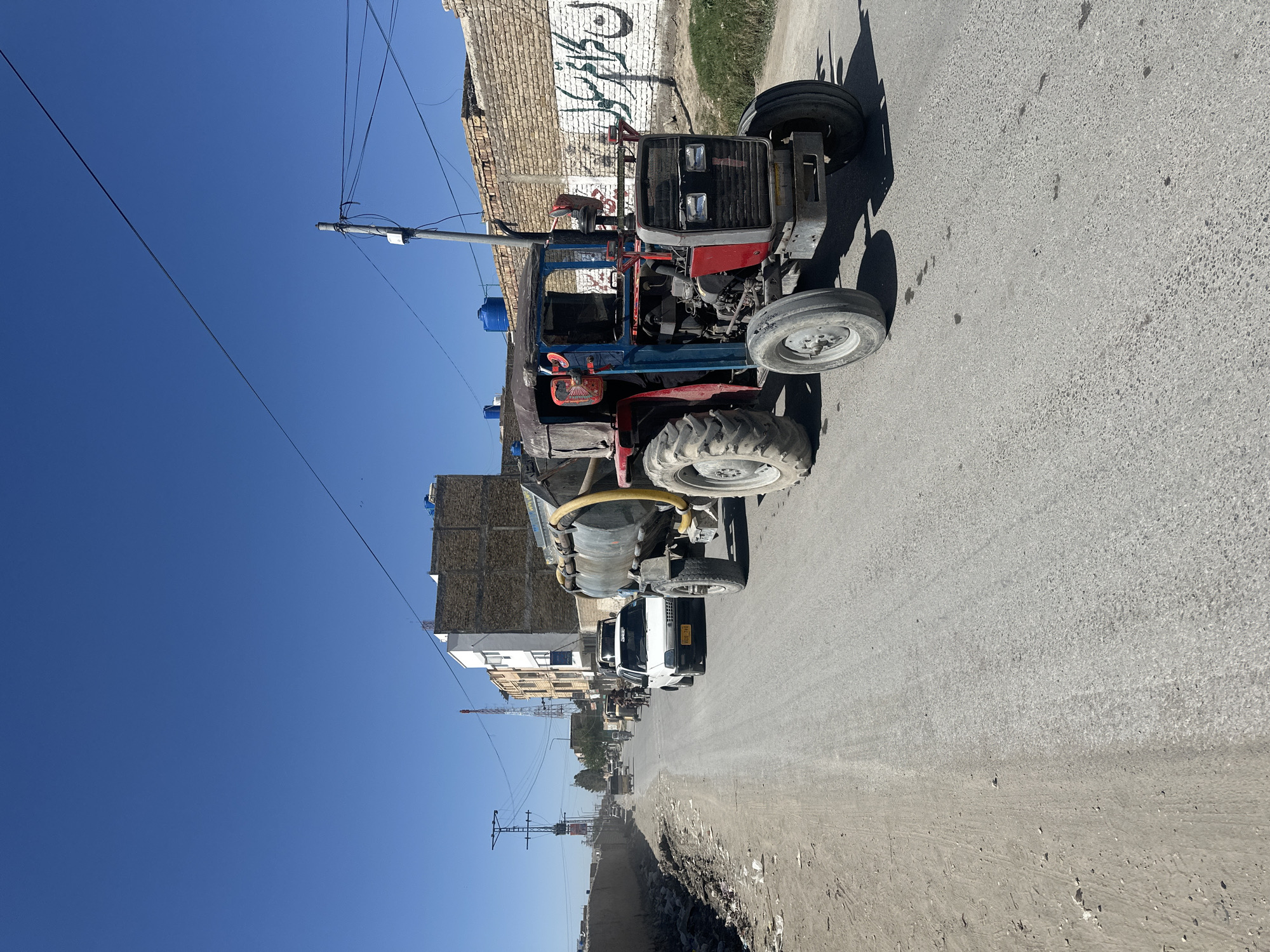 The image depicts a street in Quetta City Tehsil, Pakistan. A tractor is parked on the right side of the road. The road is paved and appears to be a main thoroughfare. Buildings of varying heights line the street. Debris and dirt are visible along the roadside. Overhead power lines cross the sky. The sky is clear and blue.