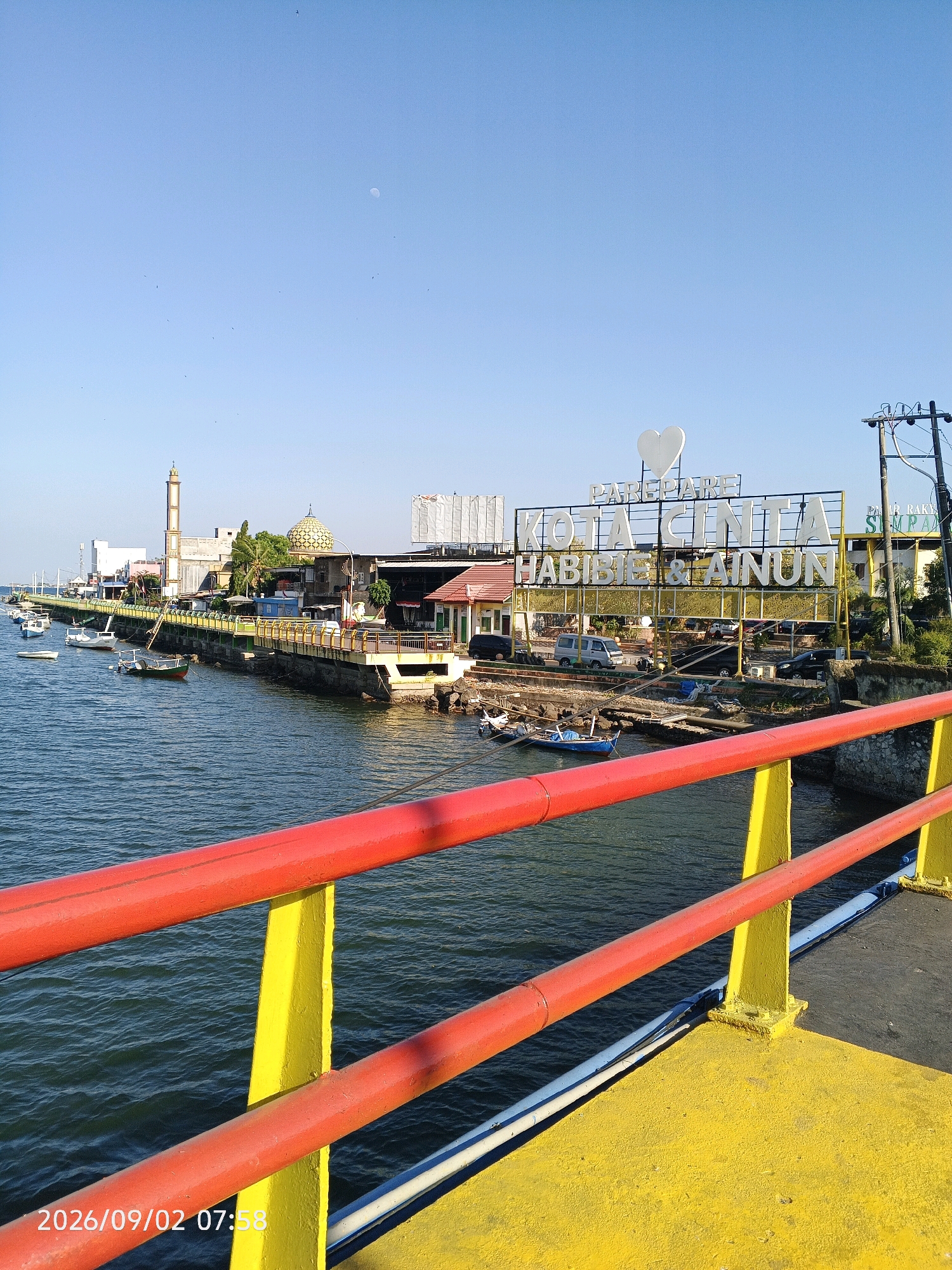 The image shows a view of the Pare-Pare waterfront in Indonesia. In the foreground, a bright yellow bridge railing with red accents runs across the bottom of the frame. The water, a deep blue, is visible below the bridge, with several small boats moored along the shore.
Along the waterfront, a yellow walkway or pier is visible, lined with a yellow railing. Buildings are clustered along this walkway, including a mosque with a golden dome and a tall minaret, and other structures that appear to be shops or restaurants. A large sign with white lettering and a white heart at the top reads "PAREPARE KOTA CINTA HABIBIE & AINUN". There are cars parked along the waterfront behind the sign.
The sky is a clear blue, with a faint crescent moon visible high up. A few birds are scattered across the sky. The overall scene depicts a sunny day in a coastal city. The timestamp on the image indicates it was taken on September 2, 2026, at 07:58.