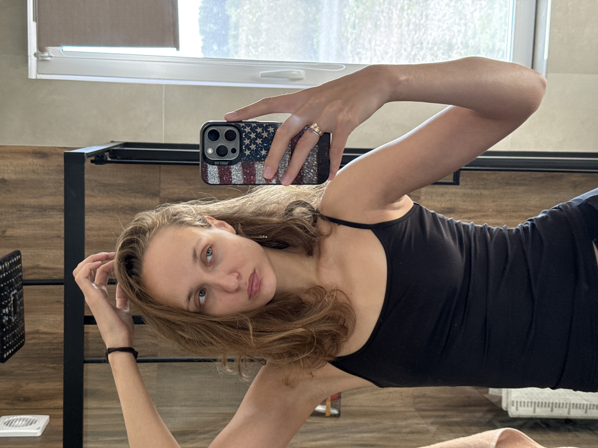 The image shows a young woman taking a selfie in a bathroom. She is standing in front of a mirror, wearing a black tank top and dark bottoms. Her right arm is raised, holding a phone with an American flag case, and her left hand is in her hair. Her expression is somewhat pouty. The bathroom has wooden-paneled walls, a showerhead visible in the background, and a window with a sheer blind. The scene is well-lit, suggesting it's daytime. There are no specific indications of the city of Подшивалово, Russia, or any other geographical markers.