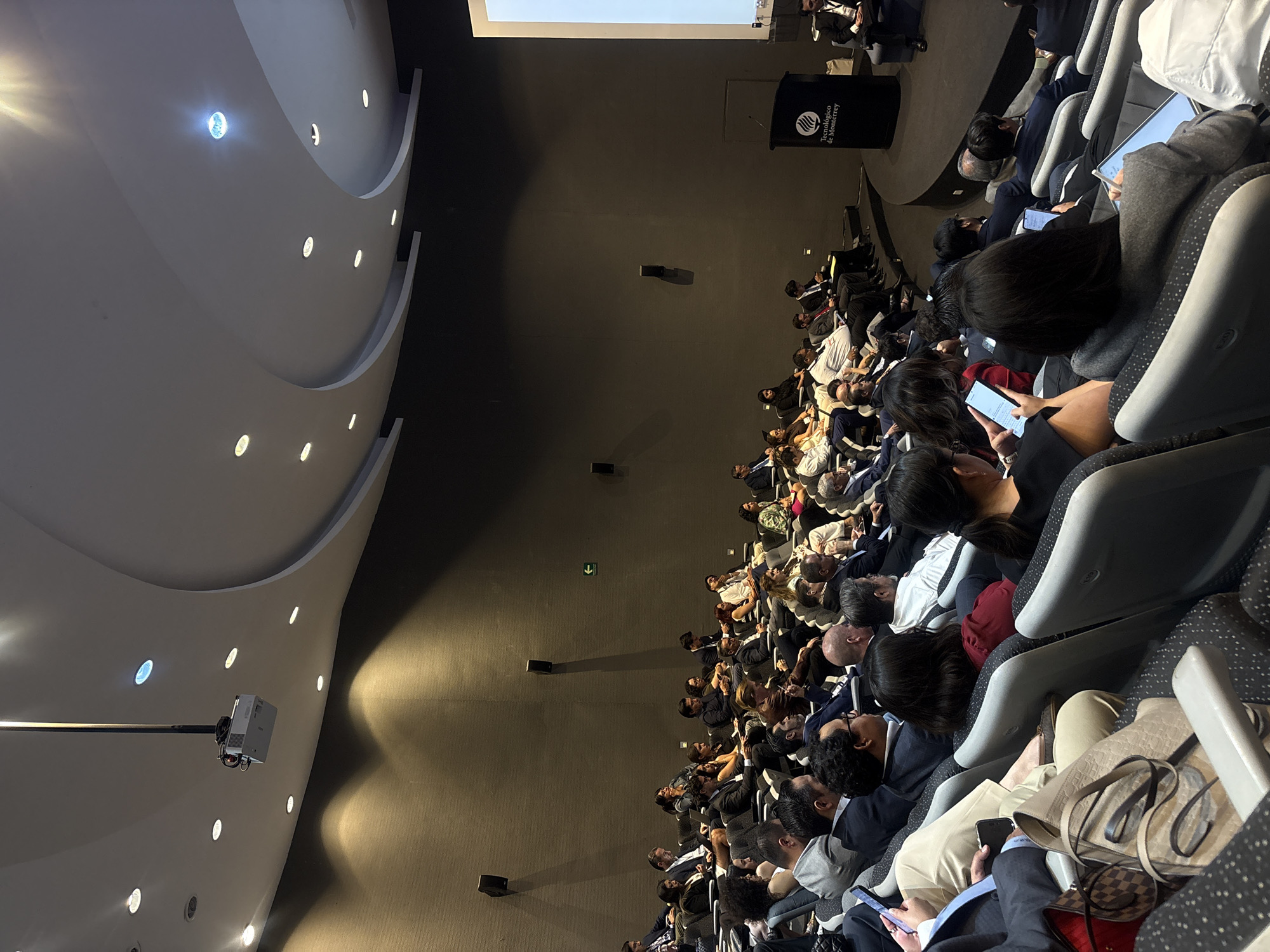 Audience members sit in rows of gray chairs in a large auditorium. Many attendees are using mobile phones. A projector hangs from the ceiling. The stage has a lectern with the logo of the "Tecnológico de Monterrey" university. The location is the city of Puebla, Mexico.