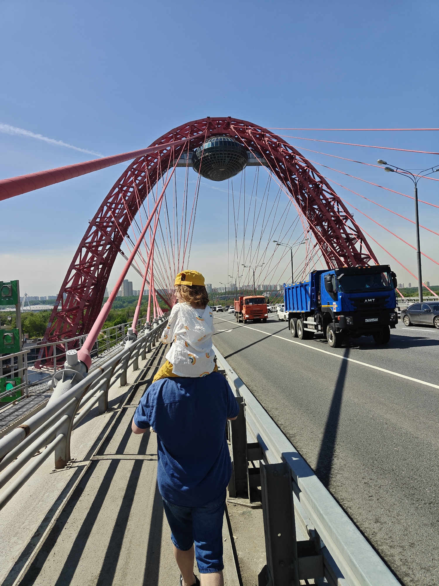 A medium shot captures a father carrying a child on his shoulders while walking along a pedestrian walkway on the Zhivopisny Bridge in Khimki, Russia. The father wears a blue short-sleeved shirt and jean shorts. The child has on a yellow hat and a white, patterned hoodie. The bridge's red arch and support cables dominate the background, and a spherical structure sits at the top of the arch. A multi-lane roadway runs adjacent to the pedestrian walkway, with several trucks and cars. Streetlights line the side of the road. In the distance, a cityscape can be seen amidst trees. The sky is a clear blue with a single white contrail. The walkway is composed of concrete with steel railings.