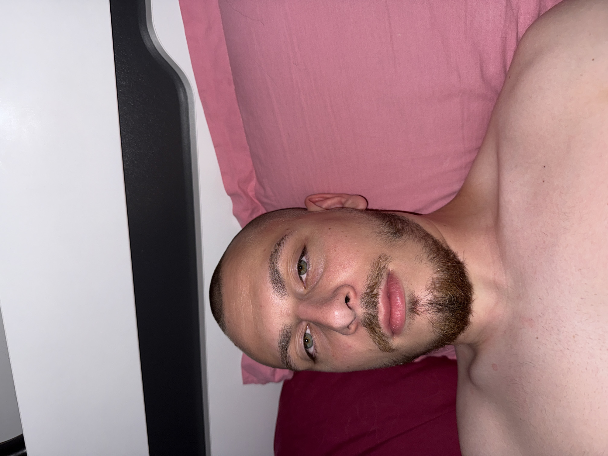 A male individual is depicted in a close-up portrait, lying in an indoor setting. The subject has fair skin, a closely shaved head, and a neatly trimmed dark brown beard and mustache. His green eyes are open and directed towards the viewer, with his lips slightly parted. His bare upper chest and collarbone area are visible, showing minimal body hair and a few small marks. He rests on pink bedding, with a pink pillow partially visible behind his head to the right. In the background, above the subject's head, a white furniture surface with a matte black horizontal band and a subtle curve on the right side is visible. The scene is situated in Stara Zagora, Bulgaria.