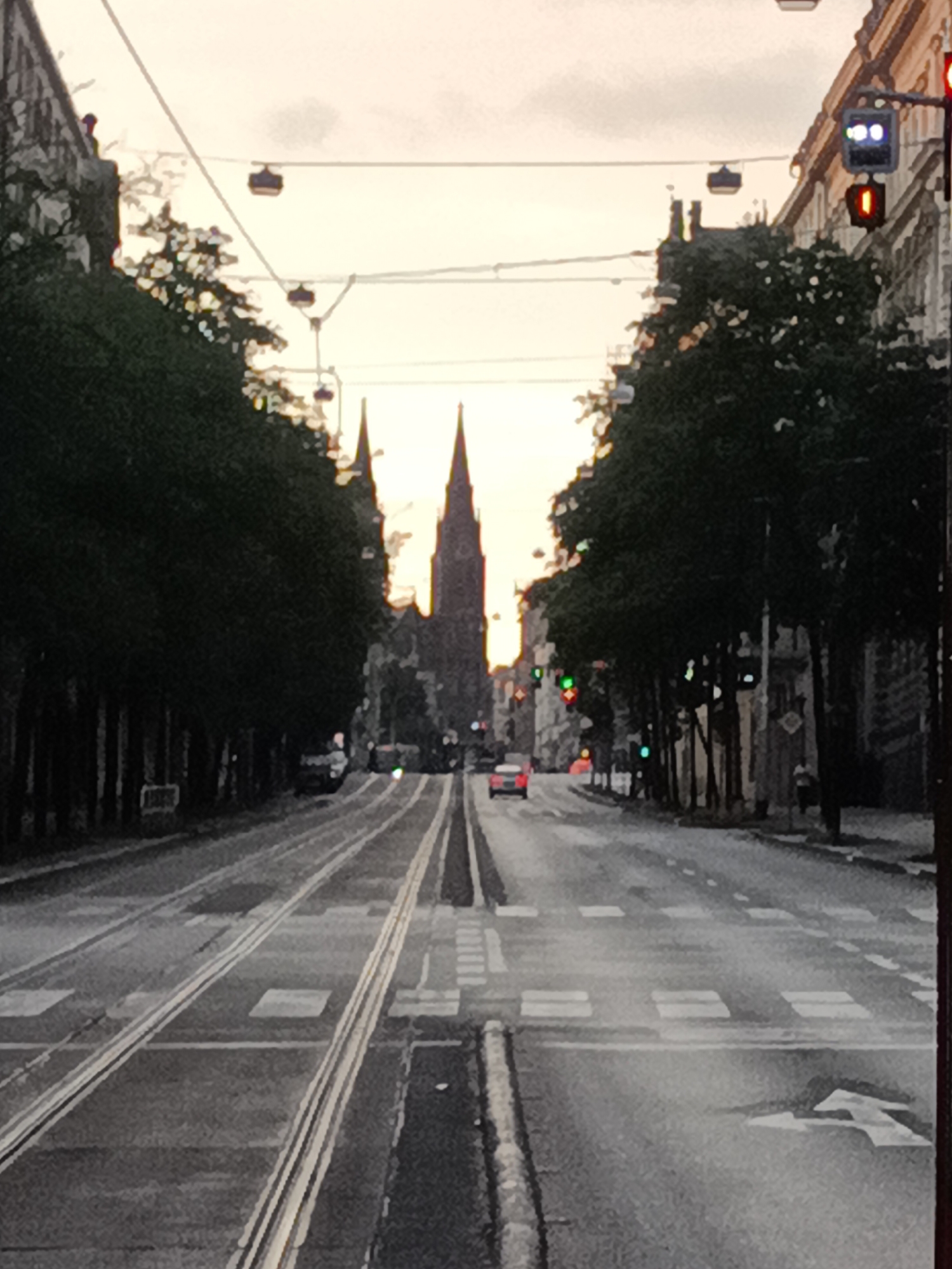 The image captures a street scene in Prague, Czechia, likely during twilight. The perspective is from a vehicle or elevated viewpoint looking down a street lined with tram tracks and trees. Two prominent church spires are visible in the distance, characteristic of Prague's architecture. Overhead, a cable car system with small cabins is suspended. Traffic lights are visible, with one showing a red light and two white lights illuminated. A red car is stopped at a traffic light, and other vehicles are implied on the road. The street is wet, suggesting recent rain. The overall atmosphere is somewhat subdued, with the sky displaying soft hues of orange and yellow.