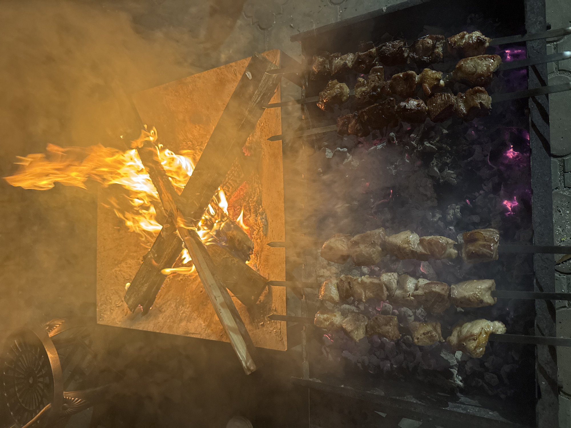 The image shows a scene with a fire and barbecue in what appears to be a backyard or outdoor cooking area. A square metal fire pit is ablaze with flames. To the right of the fire pit, metal skewers with meat cubes are arranged over a barbecue grill. Some of the meat is darker and appears more cooked than others. There is a smoky haze throughout the scene, obscuring some details. The location is possibly Tomilino, Russia, based on the prompt.