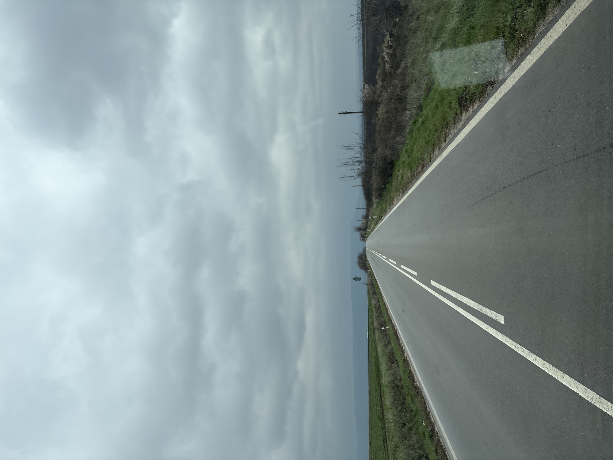The image depicts a road in Pamukchii, Bulgaria, under a cloudy sky. The asphalt road has two white dashed lines in the center and a solid white line on the right edge. The roadside vegetation is sparse, consisting of grass and shrubs. There are power lines present along the road.