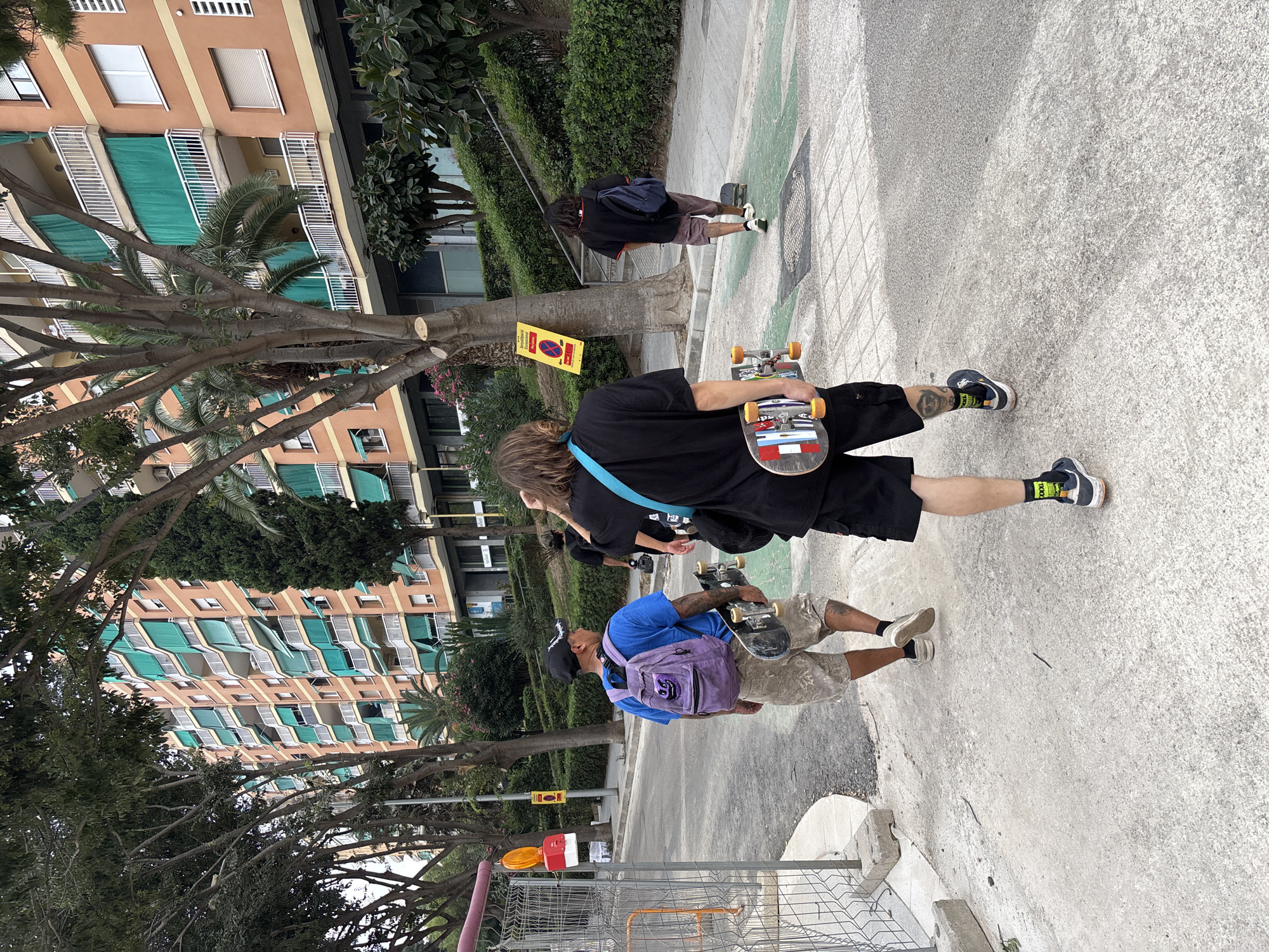 Three young men, each carrying a skateboard, walk away from the camera on a paved sidewalk adjacent to a street in Badalona, Spain. The man in the foreground wears a black t-shirt, black shorts, and black sneakers with green socks. He holds a skateboard with a colorful design. To his left, another man in a blue t-shirt and camouflage shorts carries a purple backpack and a skateboard. He wears a black baseball cap. Further down the sidewalk, a third man in a black t-shirt and maroon shorts walks away from the camera, carrying a blue backpack and a skateboard. He is partially obscured by trees. Behind the men, a large, orange apartment building with green awnings lines the street, with trees and a dense hedge in the middle ground. A yellow sign with a red prohibition symbol is posted on a pole near the curb. The scene appears to be a sunny day in an urban environment.