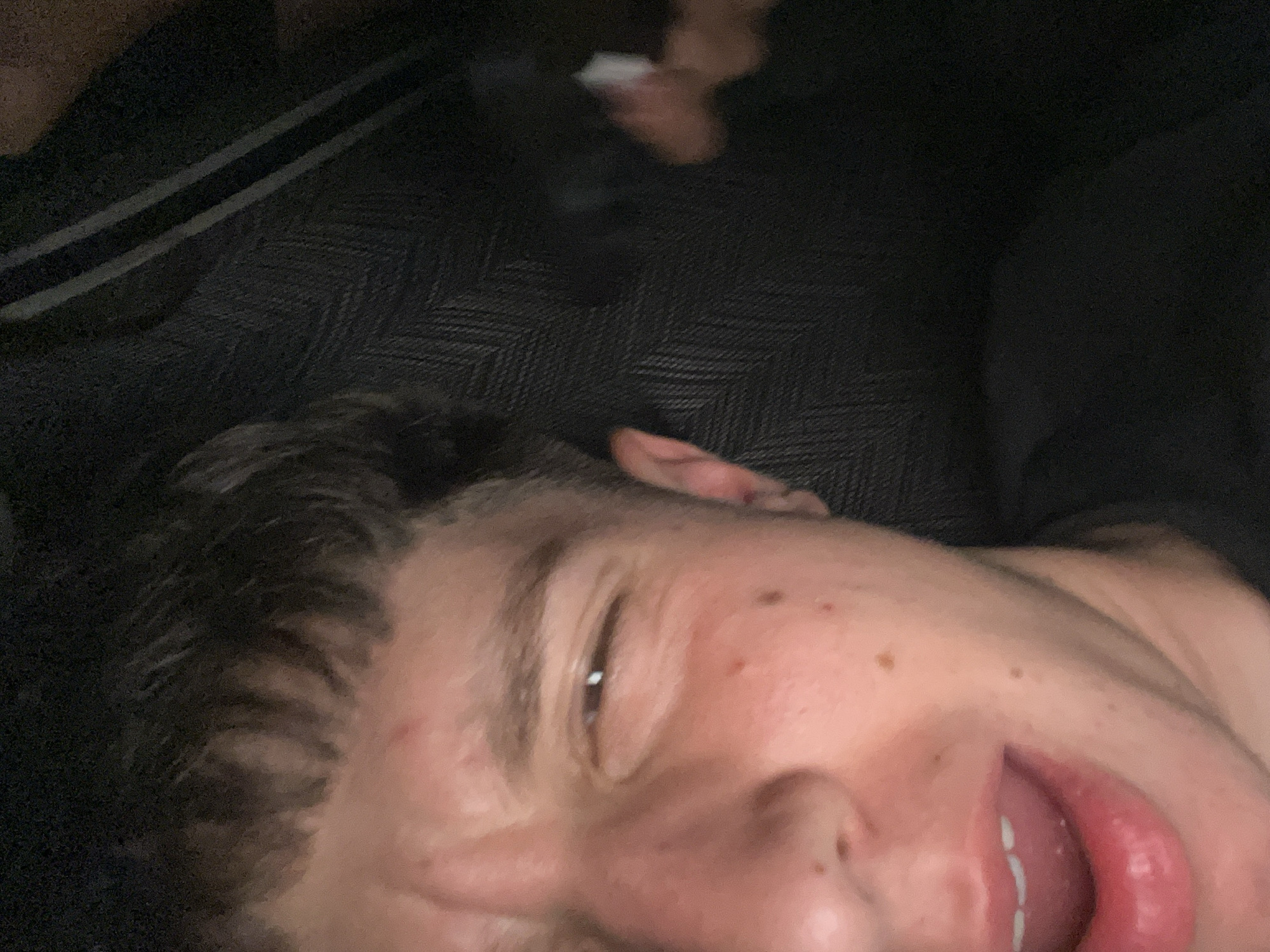 The image shows a person's face in close-up, seemingly lying down. The person has dark hair, fair skin, and is looking slightly to the side with their mouth open. The background is dark and blurred, with a patterned surface partially visible. No specific details indicate the location is Saraia, Bulgaria. No interactions or actions are discernible beyond the person's facial expression.