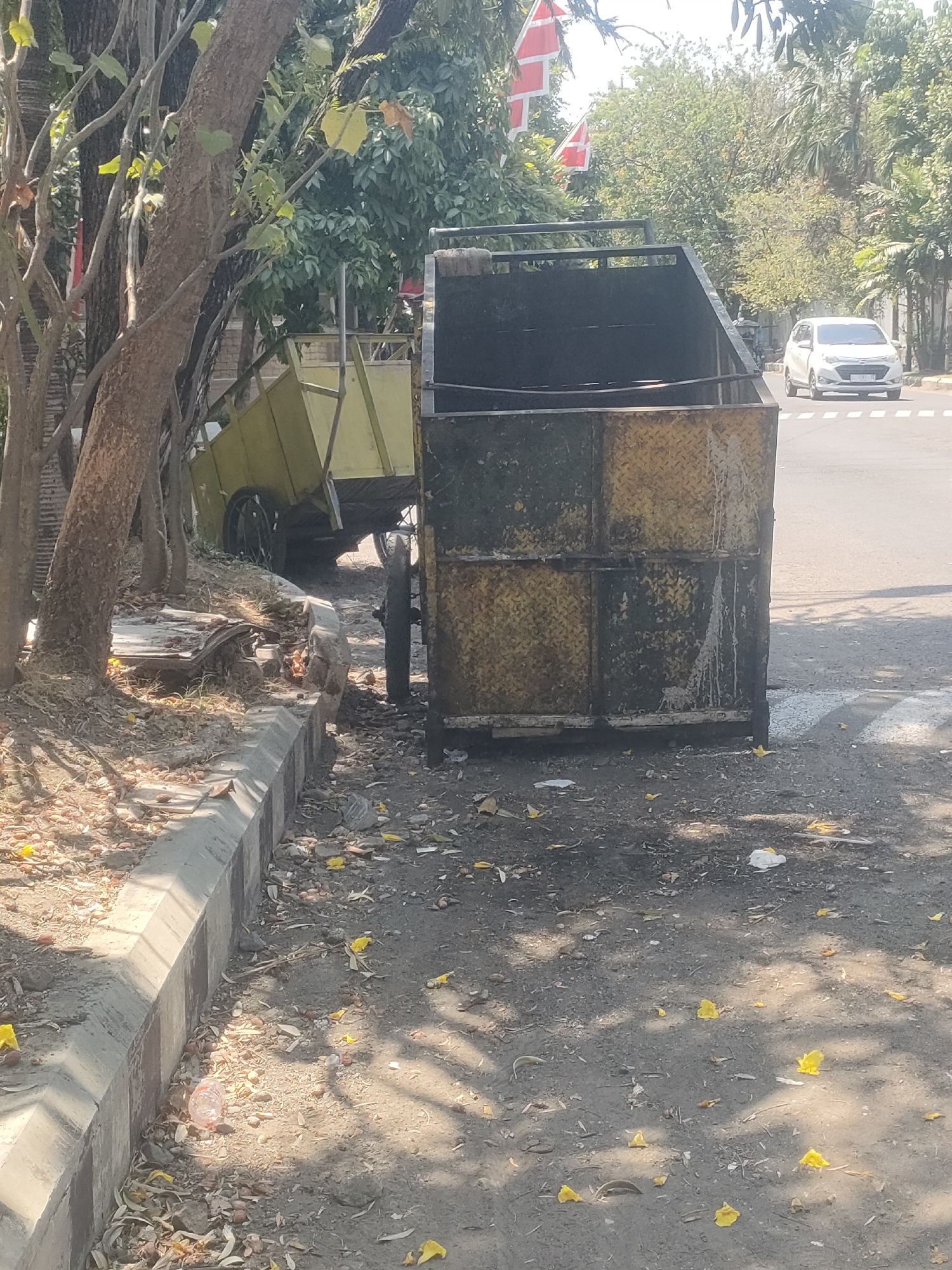 On a sunny day in Surabaya, Indonesia, an outdoor scene unfolds near a road. In the foreground, a large, industrial-looking yellow dumpster, possibly a waste receptacle, sits prominently. Its surface is textured with a diamond plate pattern and shows signs of wear and tear, including streaks of white residue. To the left of the dumpster, a smaller, overturned yellow cart with wheels is partially visible, suggesting recent activity or disarray. The ground surrounding the dumpster and cart is a mix of dirt and debris, with scattered yellow flower petals adding a touch of color. A concrete curb runs along the left side of the image, demarcating the edge of the paved area. In the background, a white car drives on the road, indicating a public thoroughfare. Trees and lush green foliage frame the scene, hinting at a well-treed urban environment. Two red and white flags, likely the Indonesian national flag, are visible flying on poles amidst the greenery, further confirming the location. The overall impression is one of an everyday street scene in Surabaya, with a focus on the utilitarian objects and natural elements present.