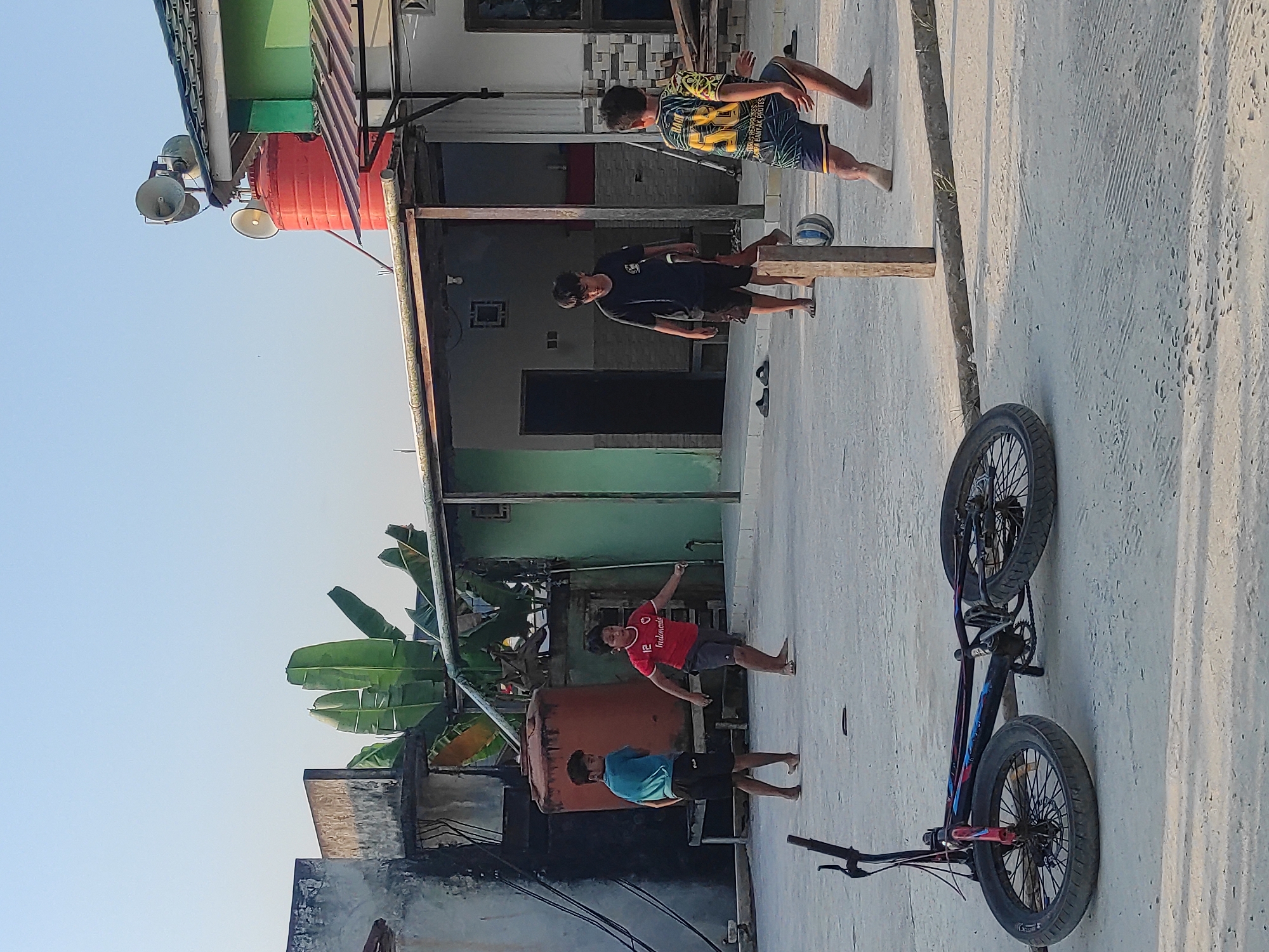 In the city of Unknown, Unknown, on a flat, dusty outdoor area, four young boys are engaged in playing soccer. The boys are dressed in casual attire, including t-shirts and shorts. Two boys are in the foreground, one wearing a blue t-shirt and black shorts, and another in a red t-shirt with the number 12 and "Indonesia" printed on it, along with gray shorts. In the background, two more boys are visible; one in a black t-shirt and dark shorts, and another in a colorful yellow and black patterned t-shirt with blue and yellow striped shorts. The soccer ball is mid-air near a wooden post.
The environment includes various structures suggesting a semi-urban or residential setting. To the left, there is a concrete building with a weathered facade and a large, orange water tank. In the background, there are more buildings with tin roofs, some painted green and white. A red water tank is visible atop one of the buildings, with large speakers mounted above it. Banana trees are also present, adding greenery to the scene. In the very foreground, a black BMX bicycle lies on its side, its wheels facing the viewer. The ground is a light-colored, rough surface, possibly concrete or packed dirt, with some debris scattered around. The sky is clear and bright blue, indicating daylight.