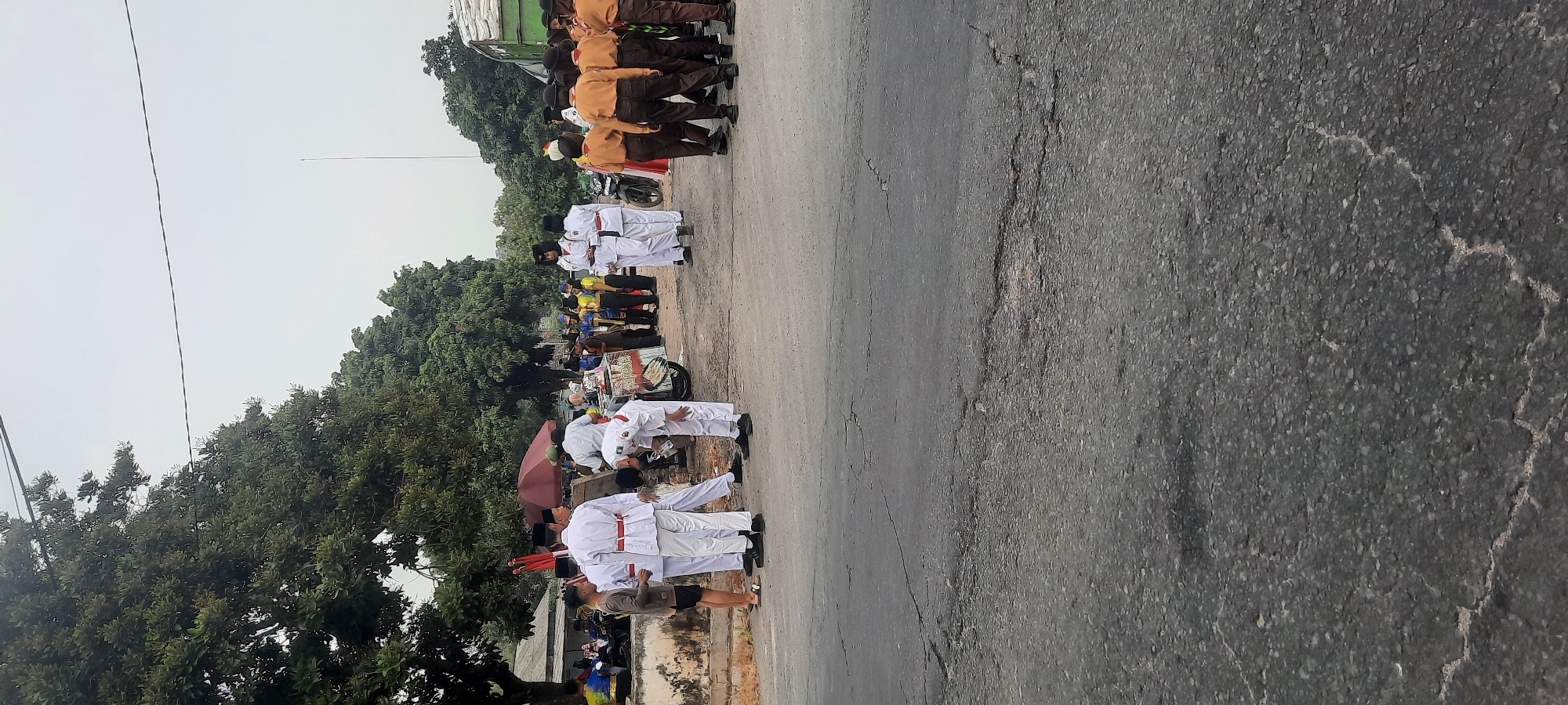 A group of individuals, many in white uniforms with red sashes, and others in brown uniforms, are gathered on a cracked asphalt road in Tangkil, Indonesia. The scene appears to be outdoors, with trees and buildings visible in the background. The sky is overcast.
In the foreground, the cracked asphalt road dominates the lower portion of the image. To the left, a few people are standing, some in white uniforms and others in civilian clothing. A young man in a brown shirt and black shorts is interacting with individuals in white uniforms. One person in a white uniform is bent over.
Further back, a cluster of people in white uniforms with red sashes are standing in a line. Behind them, a cart or vehicle with an umbrella and signage is present, and more people are gathered around it. To the right, a group of individuals in brown uniforms, possibly scout uniforms, are standing in a line. Some are looking towards the left, while others are facing forward. A motorcycle is parked behind the cart.
In the background, trees line the edge of the scene, and some low-lying buildings are visible. Power lines stretch across the upper part of the image, with a thin pole extending upwards on the right side. The overall impression is of a gathering or event taking place outdoors in what seems to be a semi-urban or rural setting.