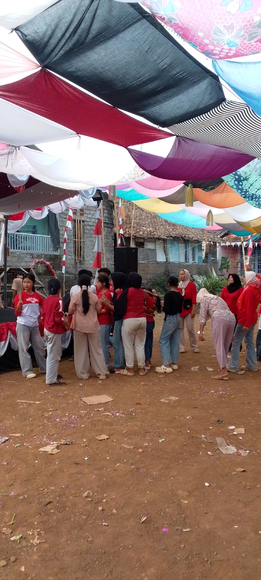 In Cilaku, Indonesia, a group of people are gathered under a canopy of colorful fabric squares. The fabrics, in shades of black, red, white, purple, and patterned designs, are stretched overhead, creating a festive, shaded area. The ground is dirt and scattered with debris. A building with a tiled roof and stone walls is visible in the background, along with a white railing and what appears to be a stage or platform decorated with red and white fabric.
Most of the individuals are wearing red tops and casual pants, suggesting a communal event or celebration. Several children are present among the adults, and their interactions appear casual and informal. The overall atmosphere seems to be one of a lively outdoor gathering.