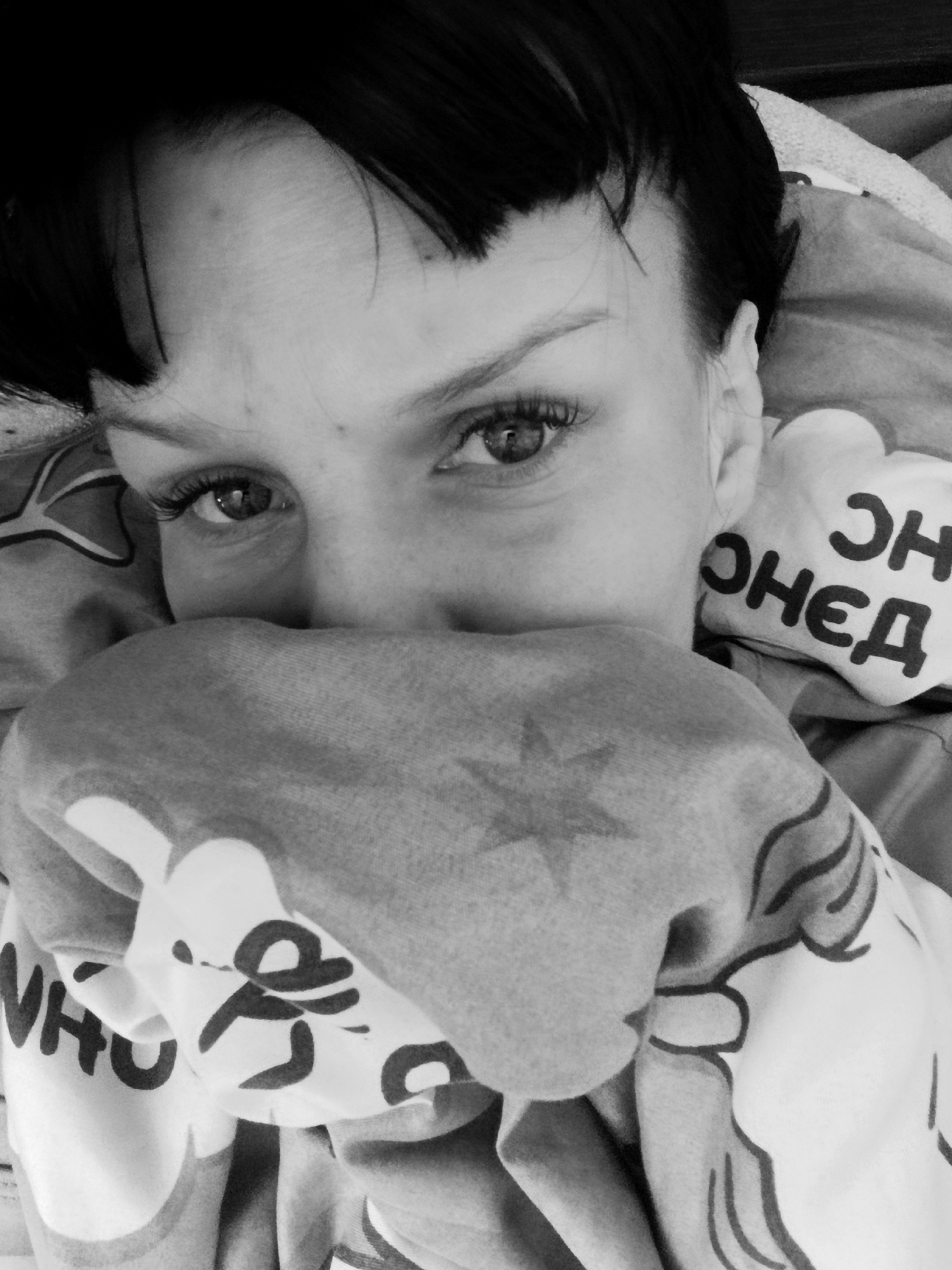A black and white, eye-level close-up shows a person with dark hair, including short bangs, partially covered by a blanket or other cloth patterned with what appears to be lettering in Cyrillic and stylized figures. The person's eyes are open and focused toward the camera. The cloth obscures the lower part of their face. Other text in Cyrillic is visible on an object next to the person’s head and reads "ОНЕД". The location is unknown.