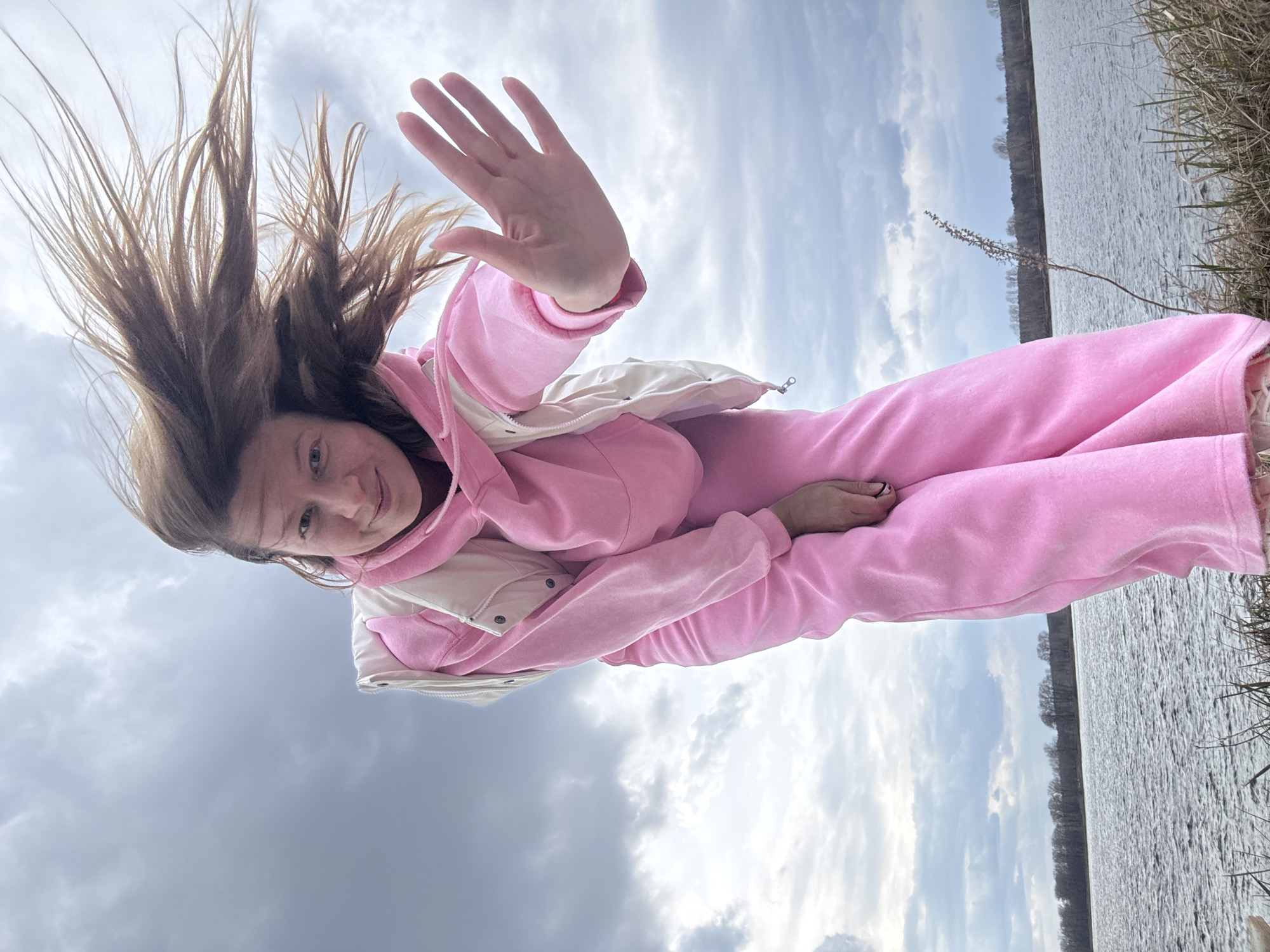 A young woman with light brown hair, appearing to be in her 20s or early 30s, is captured in a dynamic outdoor portrait. She is smiling broadly, with her head tilted slightly and her gaze directed towards the viewer. Her light brown hair is dramatically swept upwards and outwards, indicating strong wind. She is wearing a matching pink hooded sweatshirt and pink sweatpants, layered with a light-colored, possibly off-white or cream, puffy vest. Her right hand is raised, palm open, as if in a wave or gesture of greeting.
The scene is set outdoors by a large body of water, likely a lake or wide river, with a distant shoreline visible across the water. The water surface appears somewhat choppy, reflecting the cloudy sky above. The sky is predominantly overcast with varying shades of gray and white clouds, but also shows patches of lighter blue. On the immediate right side of the frame, some dry grasses or reeds are visible along the bank. In the distance, a strip of bare trees lines the far bank of the water.
The overall impression is one of a cheerful and active moment, possibly a self-portrait taken on a windy day. The location is Chaikovsky, Russia. There is no visible text in the image.
