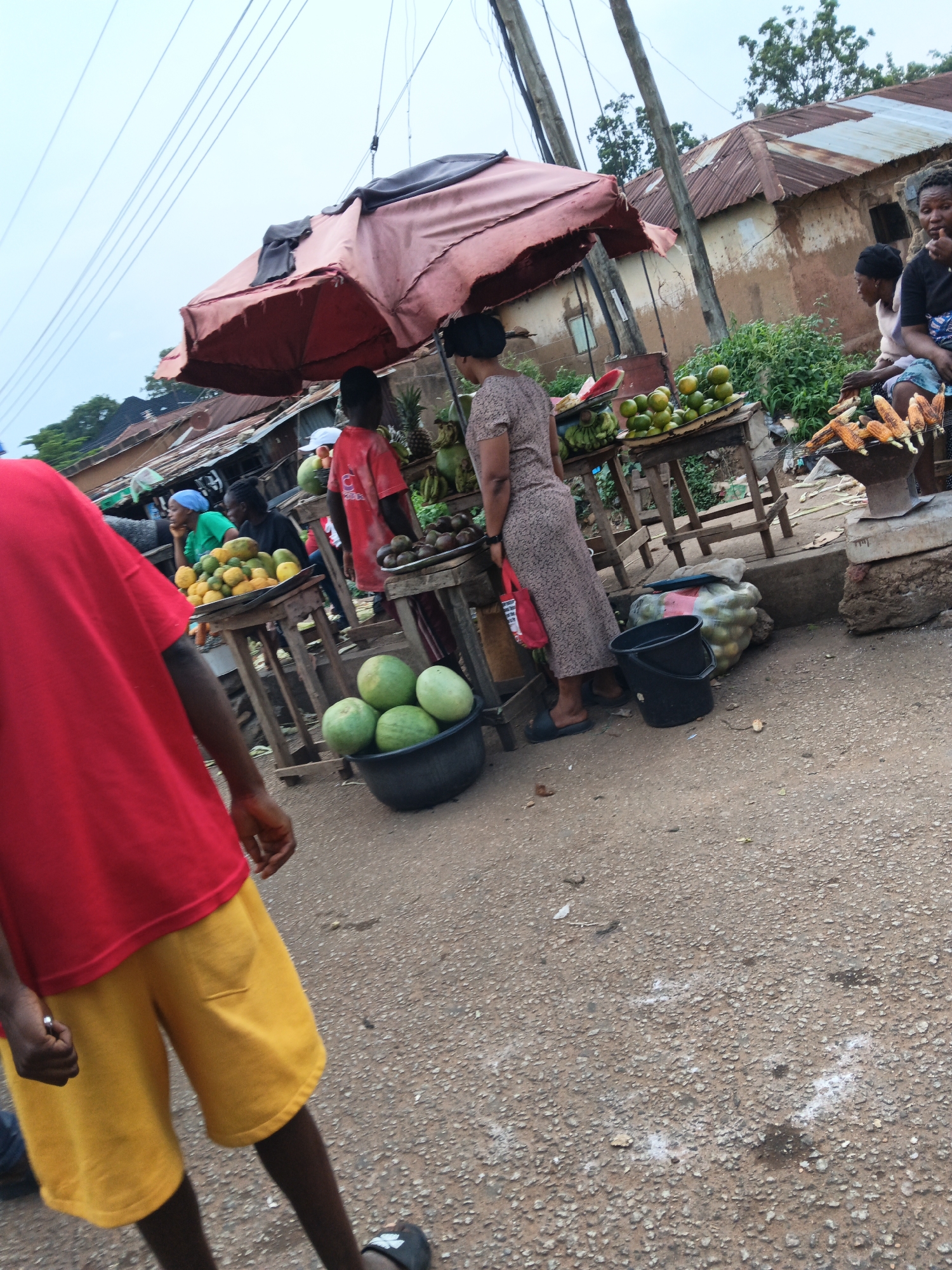 The image depicts an outdoor market scene in a location identified as Unknown, Unknown. In the foreground, a person wearing a red t-shirt and yellow shorts is partially visible, facing away from the camera. Behind them, a market stall is set up with an umbrella providing shade. Various fruits are displayed on tables, including what appear to be bananas, oranges, and possibly pineapples. Several people are present, some tending to the stalls and others browsing. One woman in a patterned dress is standing by a table of fruit. Another person, possibly a child, is wearing a red shirt and a white cap. In the background, there are buildings with corrugated metal roofs and mud walls, suggesting a rural or semi-urban setting. There are also power lines visible overhead. A black bucket is placed on the ground near the fruit stall. Roasted corn on the cob is being displayed on a grill in the mid-ground. The ground is a rough, gravelly surface.