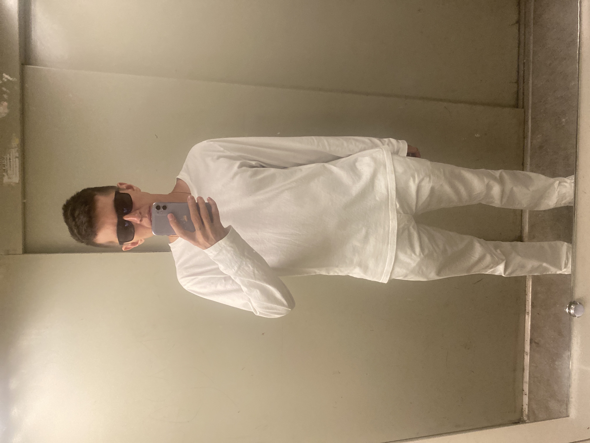 A young man wearing sunglasses, a white long-sleeved shirt, and white pants stands in what appears to be an elevator, taking a mirror selfie with a phone. The walls of the elevator are a neutral, utilitarian color. The lighting is functional, typical of an elevator interior. There is no indication of the specific location within Novokuybyshevsk, Russia.
