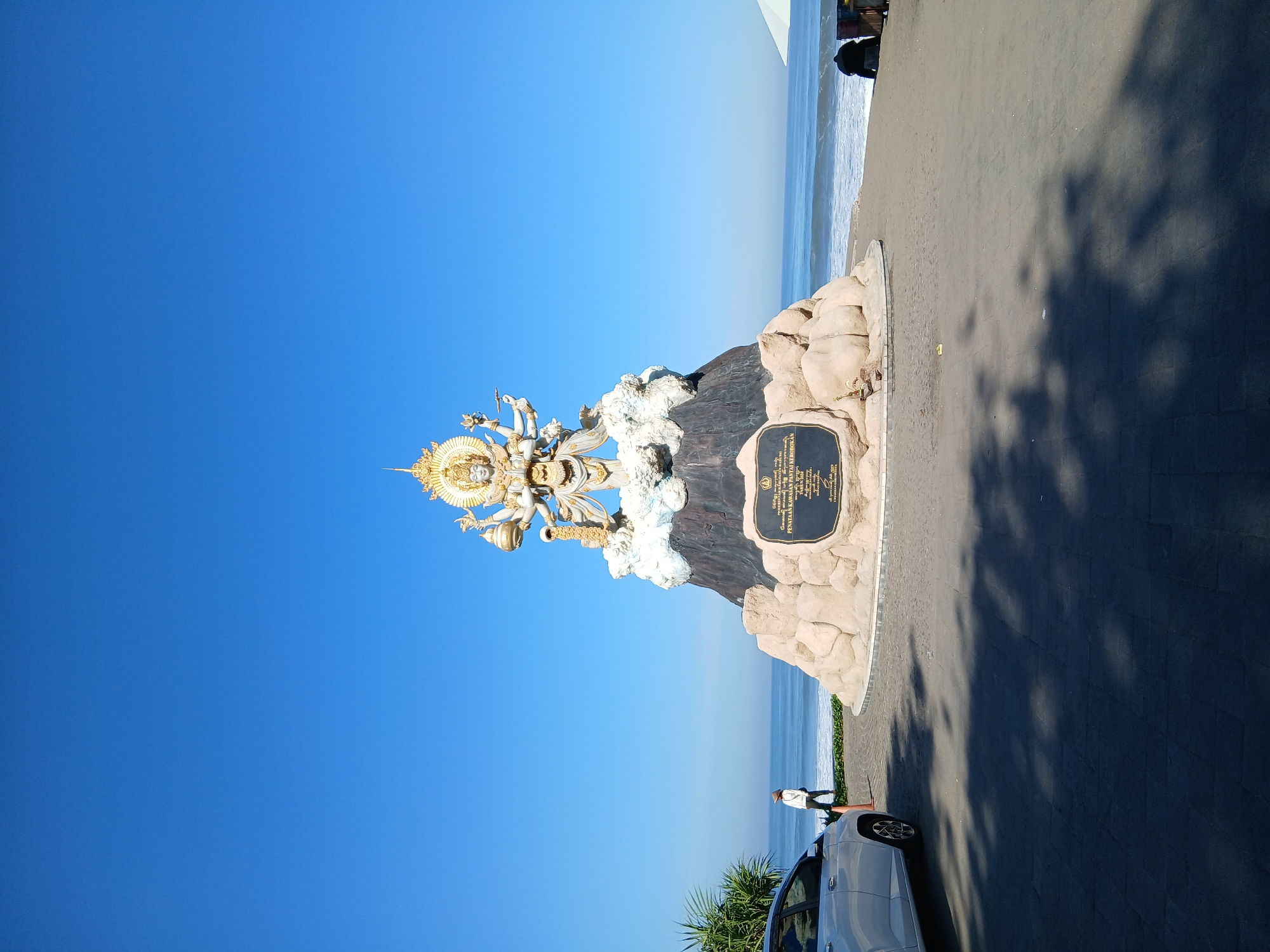 A golden statue of a multi-armed deity stands atop a rock formation on a beach in a coastal area of Indonesia. The statue is white and gold, with intricate details and a radiant halo. The rock formation is textured and sandy-colored, with some cloud-like elements around the base of the statue. A dark plaque is affixed to the front of the rock formation, bearing inscriptions.
In the foreground, a silver car is parked on a paved area, casting a shadow. A person wearing a hat and light-colored clothing stands near the car, facing the ocean. The ocean is visible in the background, with gentle waves rolling onto a sandy beach. A few beach chairs and an umbrella are present further down the beach. The sky is a clear, bright blue. The scene is set in an outdoor environment with the sea and sky as prominent elements.