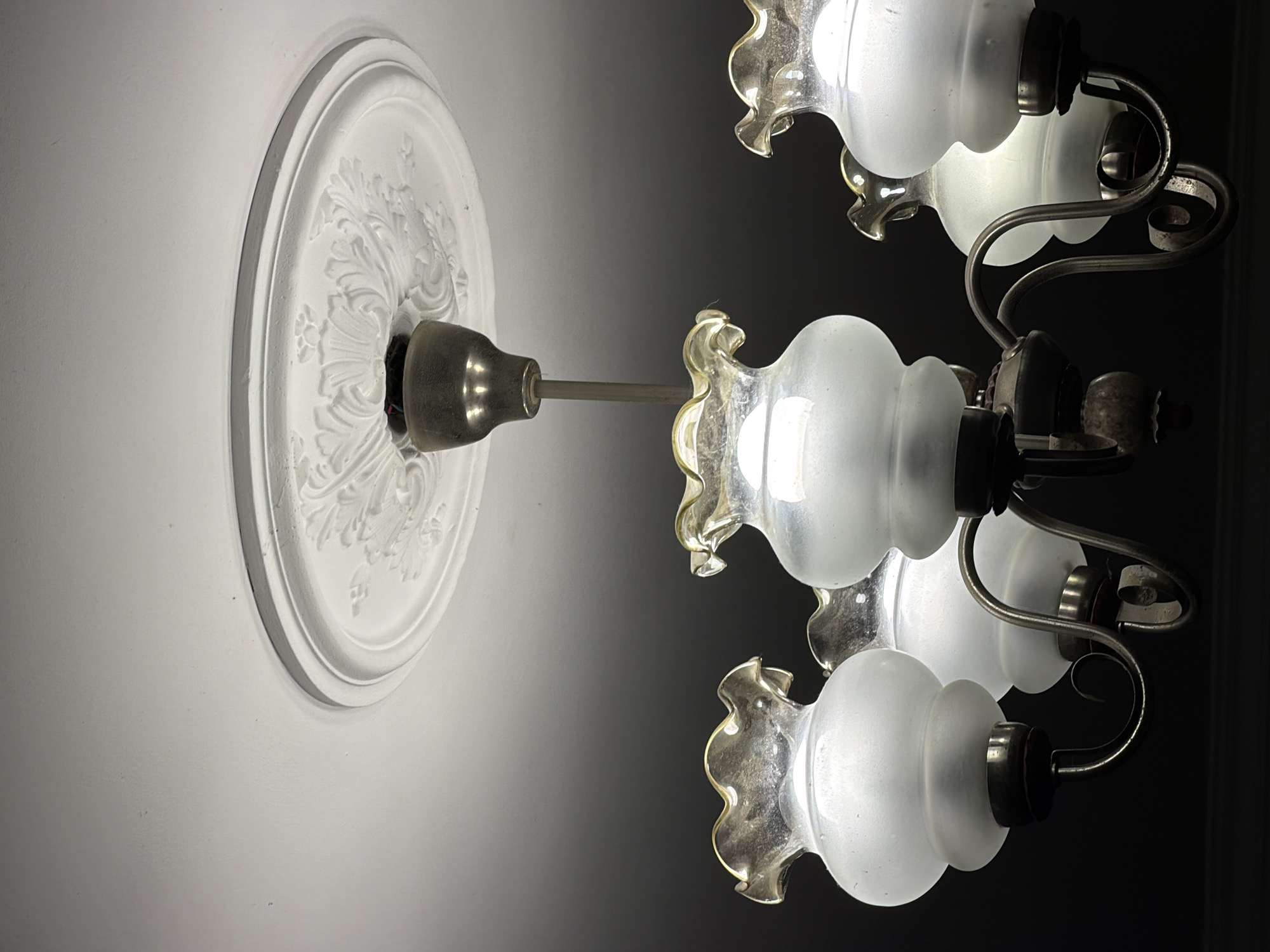 An ornate, white ceiling medallion features intricate floral detailing. A central silver fixture hangs from the medallion, supporting a chandelier with five frosted glass lampshades. Each lampshade has a ruffled, amber-tinted rim and a bulbous, frosted glass body. The chandelier arms are made of dark metal with curved designs. The background is a gradient of dark gray to black.