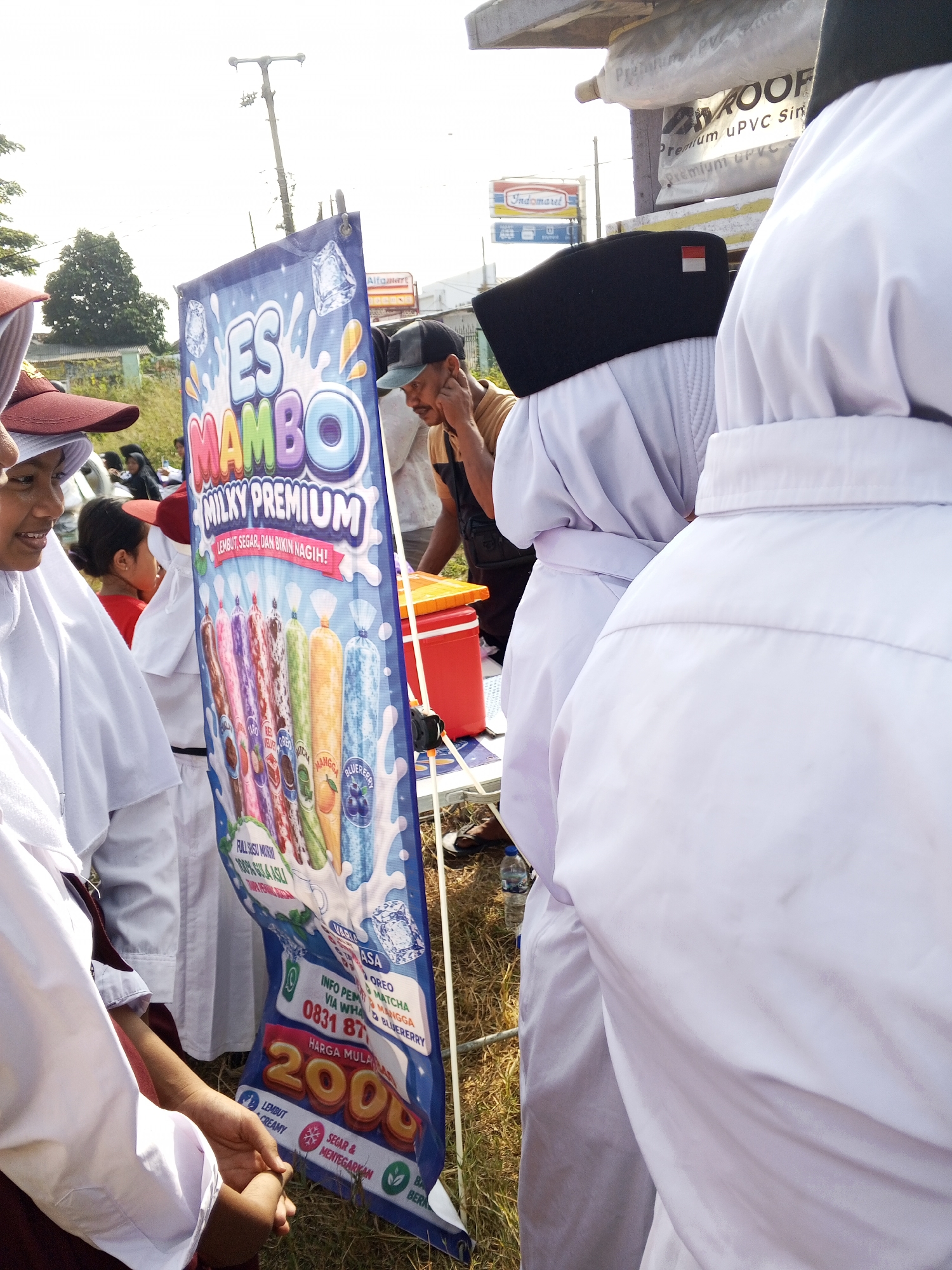 A vertical banner for "ES MAMBO MILKY PREMIUM" ice pops is displayed outdoors on a sunny day, likely in Ranjeng, Indonesia. The banner is blue and features images of ice pops in various flavors. Text on the banner includes "LEMBUT, SEGAR, DAN BIKIN NAGIH!" (Soft, Fresh, and Addictive!), "FULL SUSU MURNI 100% GULA ASLI" (100% Pure Milk, 100% Real Sugar), flavor names like "OREO", "MANGO", "MATCHA", and "BLUEBERRY", contact information "INFO PEMESANAN VIA WHATSAPP 0831 8793 6777", and "HARGA MULAI 2000" (Price Starts from 2000).
Several individuals are present. In the foreground, on the left, two people are partially visible, wearing white robes and red headscarves. One of them, a young girl, is looking to her left and smiling. Behind them, a man in a brown shirt and a black cap is visible, holding his hand to his face. Further back, a person in a black cap with a small Indonesian flag on it is visible. In the background, there are signs for "Alfamart" and "Indomaret", indicating a commercial area. A red cooler is placed on the ground next to the banner. The ground is covered in dry grass, and the sky is bright and overcast. The overall scene suggests a street vendor or outdoor event selling ice cream.