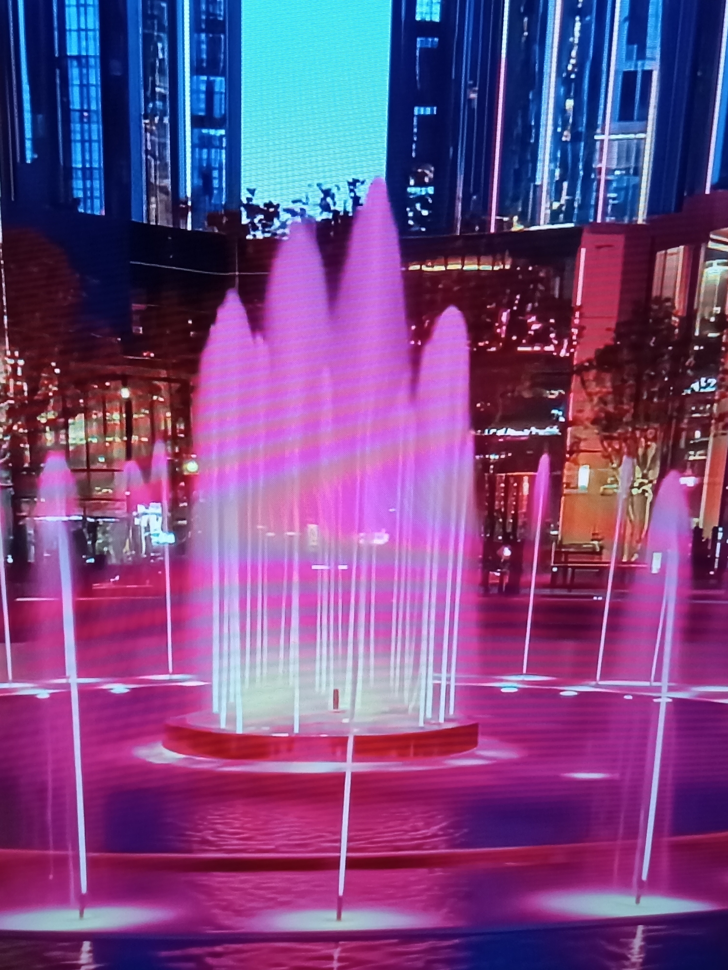 The image depicts a contemporary fountain display at night, likely in an urban setting. The fountain jets are illuminated with vibrant pink and purple lights, creating a striking visual effect against the dark surroundings. In the background, towering buildings with illuminated windows are visible, suggesting a city skyline. The architecture appears modern and sleek, with prominent vertical lines and glass facades. The water in the fountain is a deep magenta color, reflecting the colorful lights. The overall scene conveys a sense of urban nightlife and modern aesthetic. The image does not provide any specific information about the location in the city of Brno, Czechia.