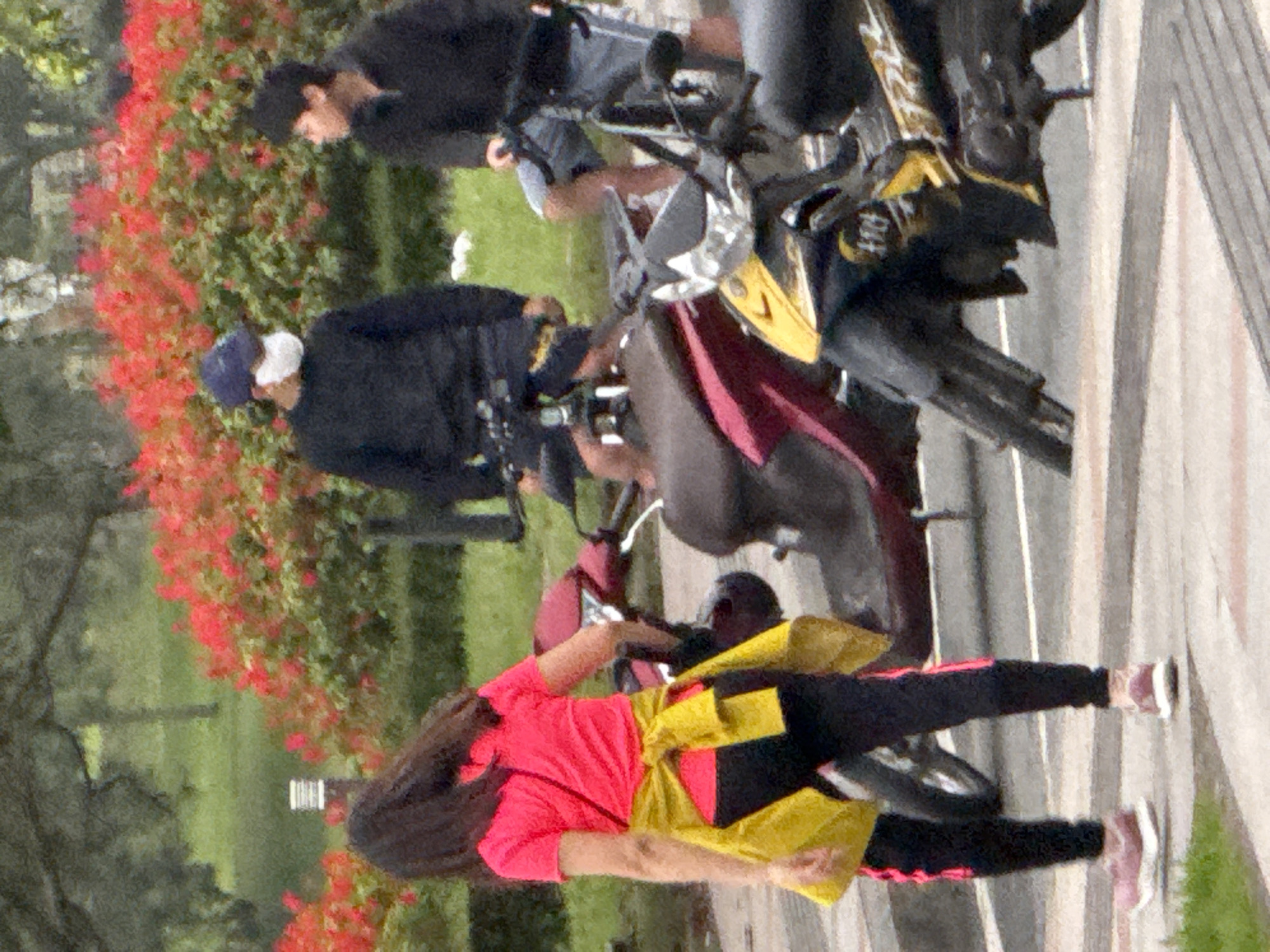 In Xuân Hương Lake, Đà Lạt, Vietnam, three individuals are captured in a scene featuring multiple motorcycles. A young girl in a bright pink t-shirt and black leggings with pink stripes stands in the foreground, a yellow sash tied around her waist. To her left, a man wearing a dark sweatshirt and a blue cap with a white face mask is visible. To the right of the girl, another person in a black sweatshirt is partially obscured by a motorcycle. Two scooters are present: a maroon one behind the girl and a black and yellow one in the foreground. The environment includes lush green grass and vibrant red flowering bushes in the background.