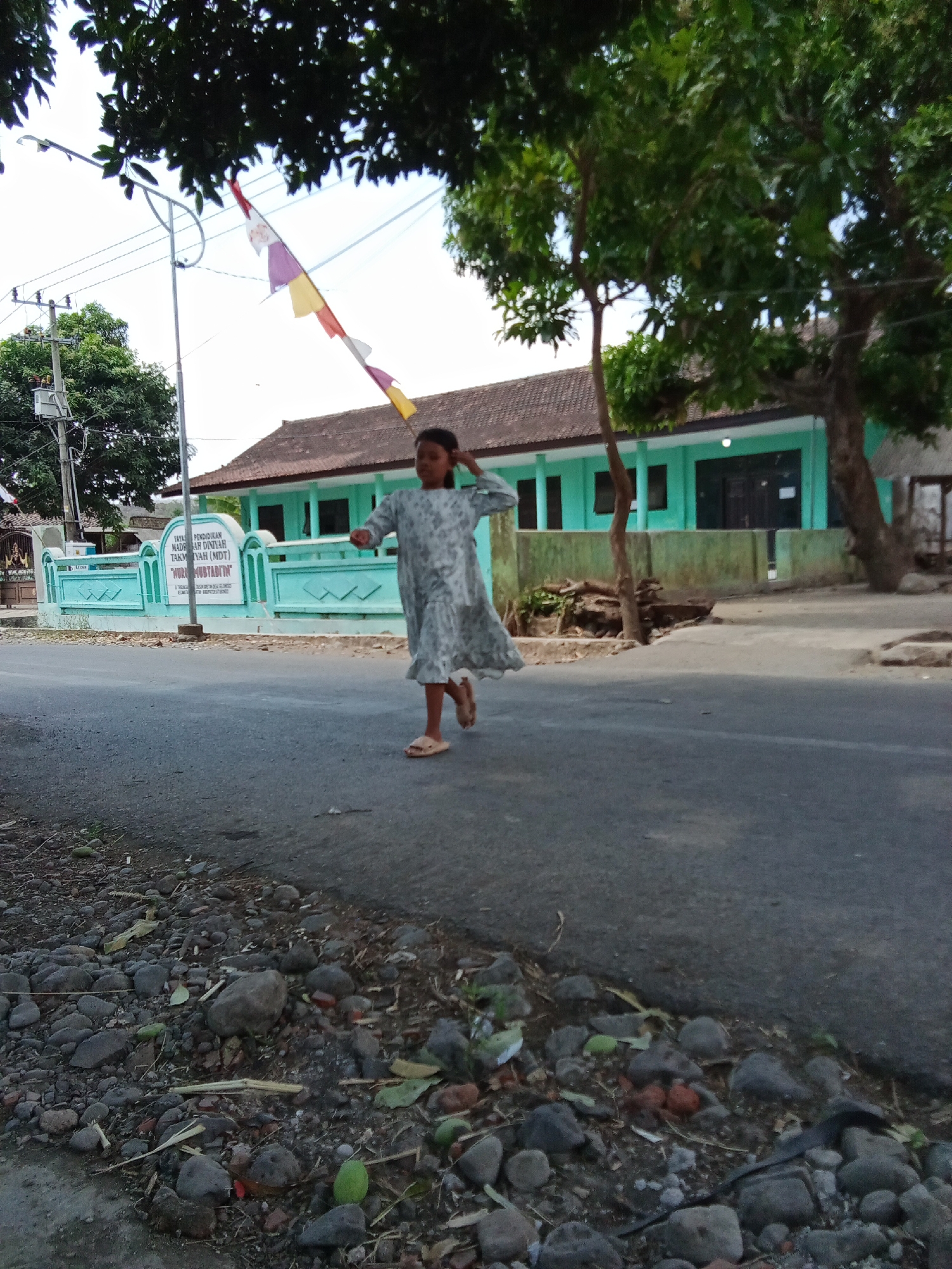 A young girl in a grey and white patterned dress runs on an asphalt road in front of a teal building with a tiled roof. She is barefoot and appears to be mid-stride, with her right arm raised and her head turned slightly to her left. The building has multiple windows and doors and a fence in front of it. A flagpole with colorful triangular flags stands to the left of the building. Trees with green leaves are visible behind and to the right of the building. The ground in the foreground is covered with rocks and debris. The sky is bright and overcast. This scene is located in Trebungan, Indonesia.