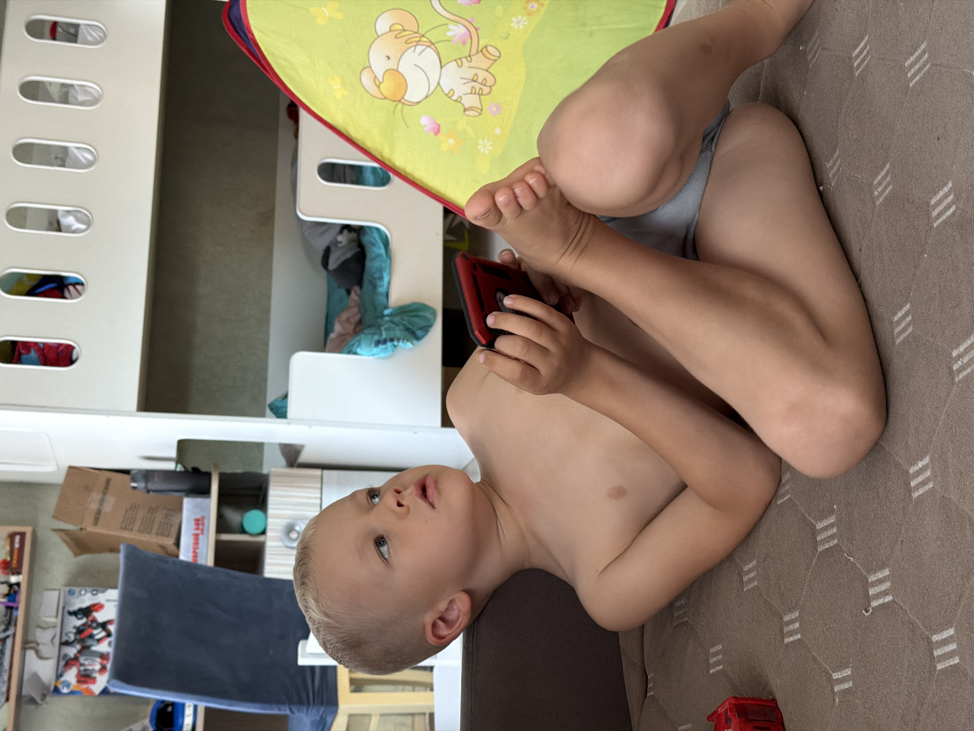 The image, taken in Sochi, Russia, features a young child lying on their back on a light brown, textured sofa or mattress. The child has short, light blond hair and light blue eyes, looking upwards and to the right with a thoughtful or focused expression. They are shirtless and wear light blue briefs. In their hands, they hold a red portable electronic device, likely a gaming console or smartphone.
Above the child's head, their feet are visible, resting on a green fabric surface that features a cartoon tiger (resembling Tigger) and small pink and yellow flowers. This fabric appears to be part of a bed or play mat. To the left, a white bunk bed frame with multiple oval cutouts is prominent.
The background shows an indoor setting, possibly a child's room. Visible through and around the bunk bed structure are shelves, a brown cardboard box, and a dark blue cushion or chair. A small red toy truck is present on the sofa near the bottom right of the frame. The ambient light suggests it is daytime. No discernible text is clearly visible on any objects in the image.