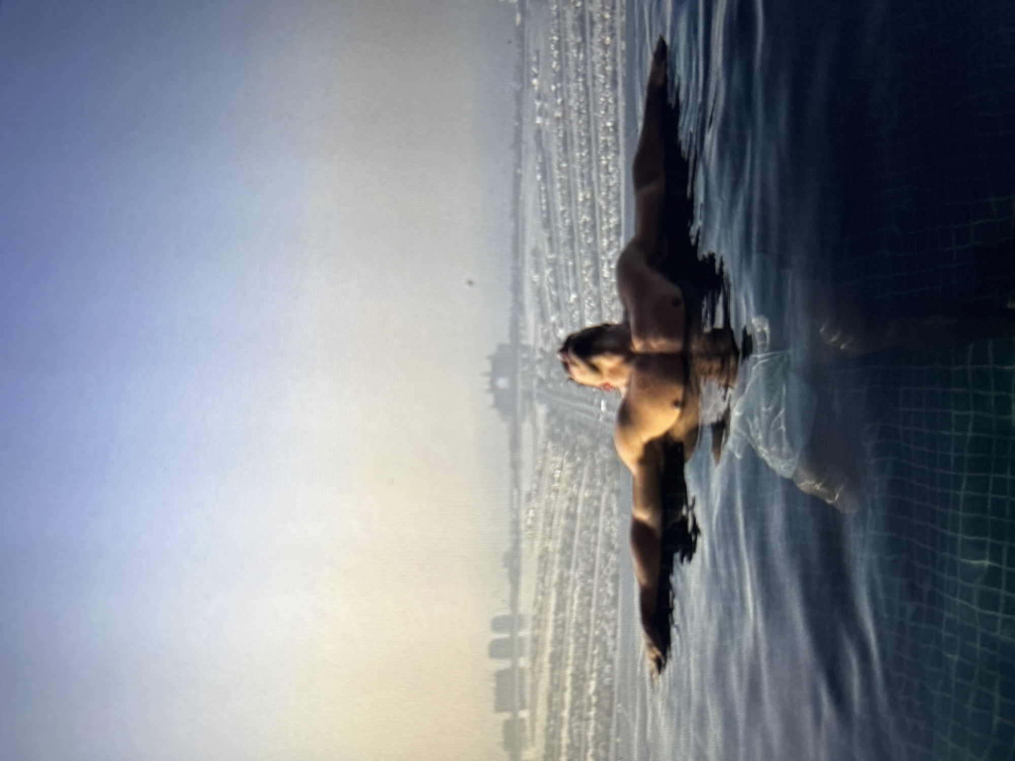 In the foreground, a man with a muscular physique is partially submerged in a swimming pool, his arms outstretched and resting on the edge. The water is clear and dark blue, revealing the tiled bottom of the pool. The man's head is tilted back, looking towards the sky.
In the background, a hazy cityscape with buildings is visible. The scene appears to be an urban environment, possibly a city by the sea, given the vast expanse of water and the distant structures. The lighting suggests it is either early morning or late afternoon, with a soft, diffused light casting a warm glow over the scene. The overall impression is one of relaxation and tranquility.
The location provided in the prompt, Burgas, Bulgaria, is not directly discernible from the visual information in the image. Therefore, I cannot confirm or deny its presence.