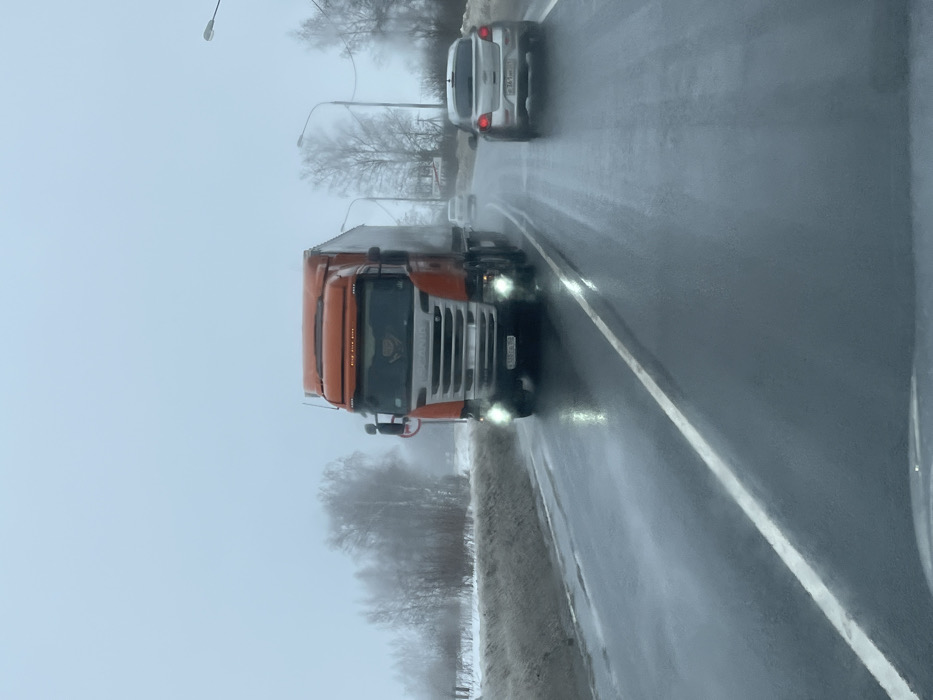 The image depicts a two-lane asphalt road in Pokrovskoe, Tver Oblast, Russia, under overcast and hazy winter conditions. The road surface is wet, indicating recent precipitation or melting snow. An orange Scania semi-truck, featuring a white box trailer and illuminated headlights, occupies the oncoming lane, moving towards the viewer. Its Russian license plate displays X538A A 69. In the same lane as the perspective of the camera, a silver Chevrolet Cruze hatchback travels away from the viewer, showing Russian license plate M 361 MH 69. A third, less distinct, light-colored vehicle is visible further ahead in the distance. The left roadside exhibits a snow-covered verge and a line of bare deciduous trees obscured by fog. The right roadside features additional bare trees and utility poles. The sky is uniformly light grey and appears foggy. No pedestrians or individuals are visible outside the vehicles.