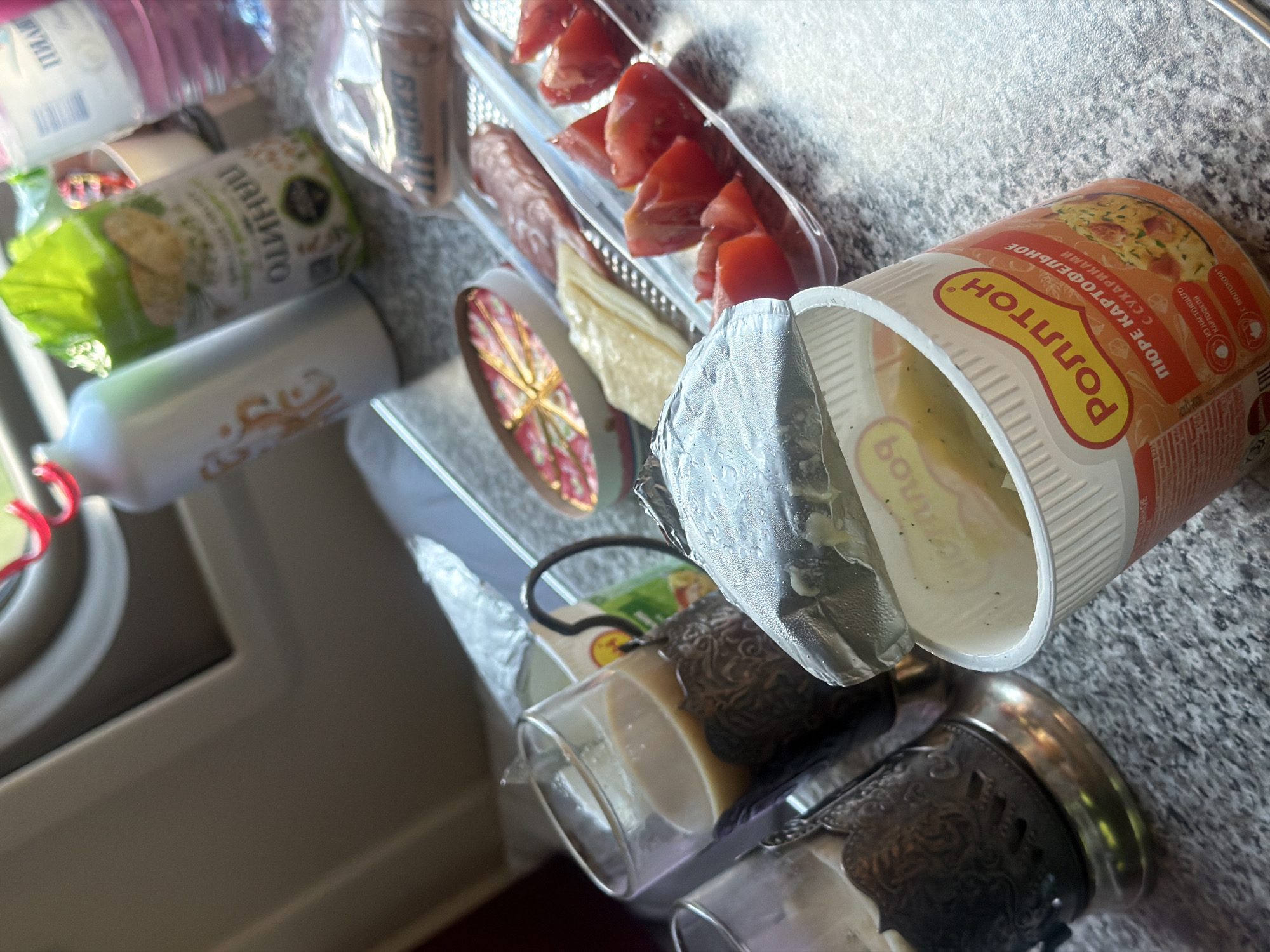 An overhead shot captures a tray of food and drinks, likely from a train or roadside establishment. A cup of instant mashed potatoes, labeled "РОЛЛТОН" and "ПЮРЕ КАРТОФЕЛЬНОЕ С СУХАРИКАМИ," is open in the foreground, with its foil lid partially peeled back. To the left, two metal cup holders contain glasses of a light-colored beverage, possibly tea or coffee. The tray also holds sliced tomatoes, a piece of cheese, and what appears to be salami. In the background, a bottle of water, a white bottle with an orange design, and a green package of snacks are visible. The overall setting suggests a travel meal, possibly in a compartment with a window showing a blurred outdoor scene. No people are present in the image.